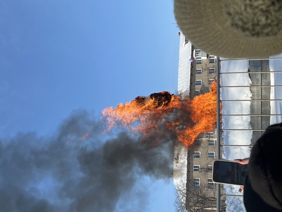 A large effigy, composed of combustible materials, is ablaze in an open area of Chaikovsky, Russia, emitting intense orange flames and a significant column of dense black smoke against a clear blue sky. The fire rises notably above a multi-story residential building situated behind it, which features a snow-covered roof and multiple windows. A purple balloon is visible, tethered to a balcony on a similar, more distant building. Snow covers the ground in the foreground and mid-ground. Bare trees are visible in the background. In the immediate foreground, a metal railing fence is present. On the left, a person with painted fingernails holds a smartphone horizontally, recording the event; the screen reflects the flames. A second observer's head, wearing a light-colored hat, is partially visible on the right.
