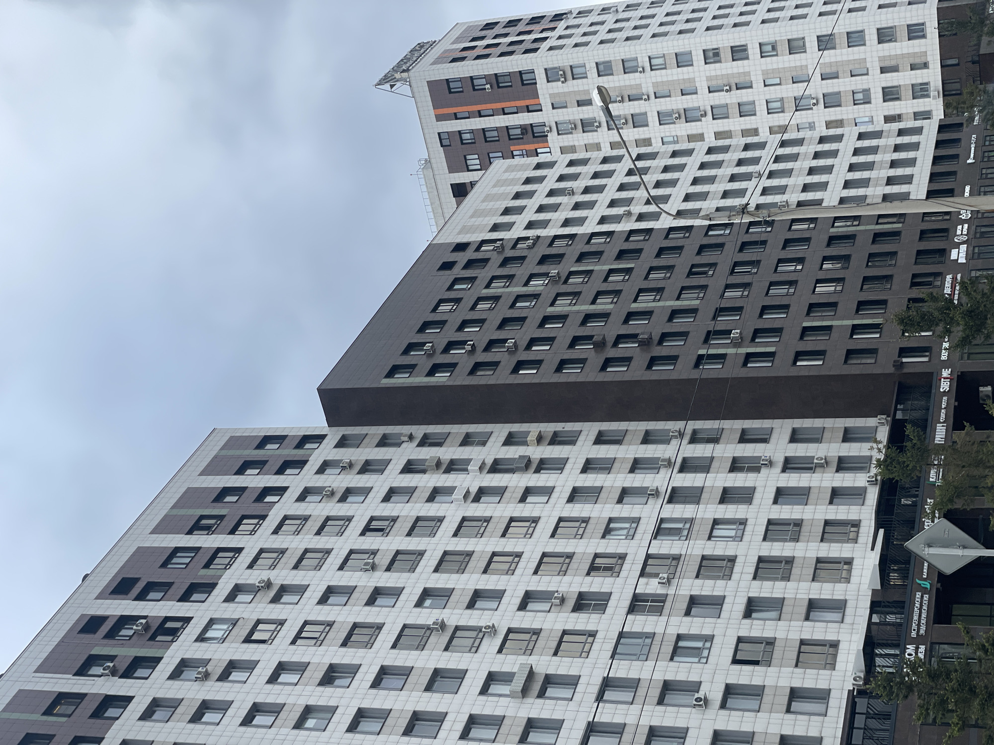 The image shows a low-angle view of a cluster of modern apartment buildings in Krasnoyarsk, Russia, notable for their repetitive grid of windows and air conditioning units. The buildings display variations in color schemes across their facades, transitioning from lighter shades at the top and bottom to darker grey or brown tones in the middle sections. The sky is overcast, and a street lamp extends into the frame, adding a vertical line. Fragments of street signs and tree foliage are visible on the right, indicating a street-level perspective. The architecture suggests a dense urban environment.