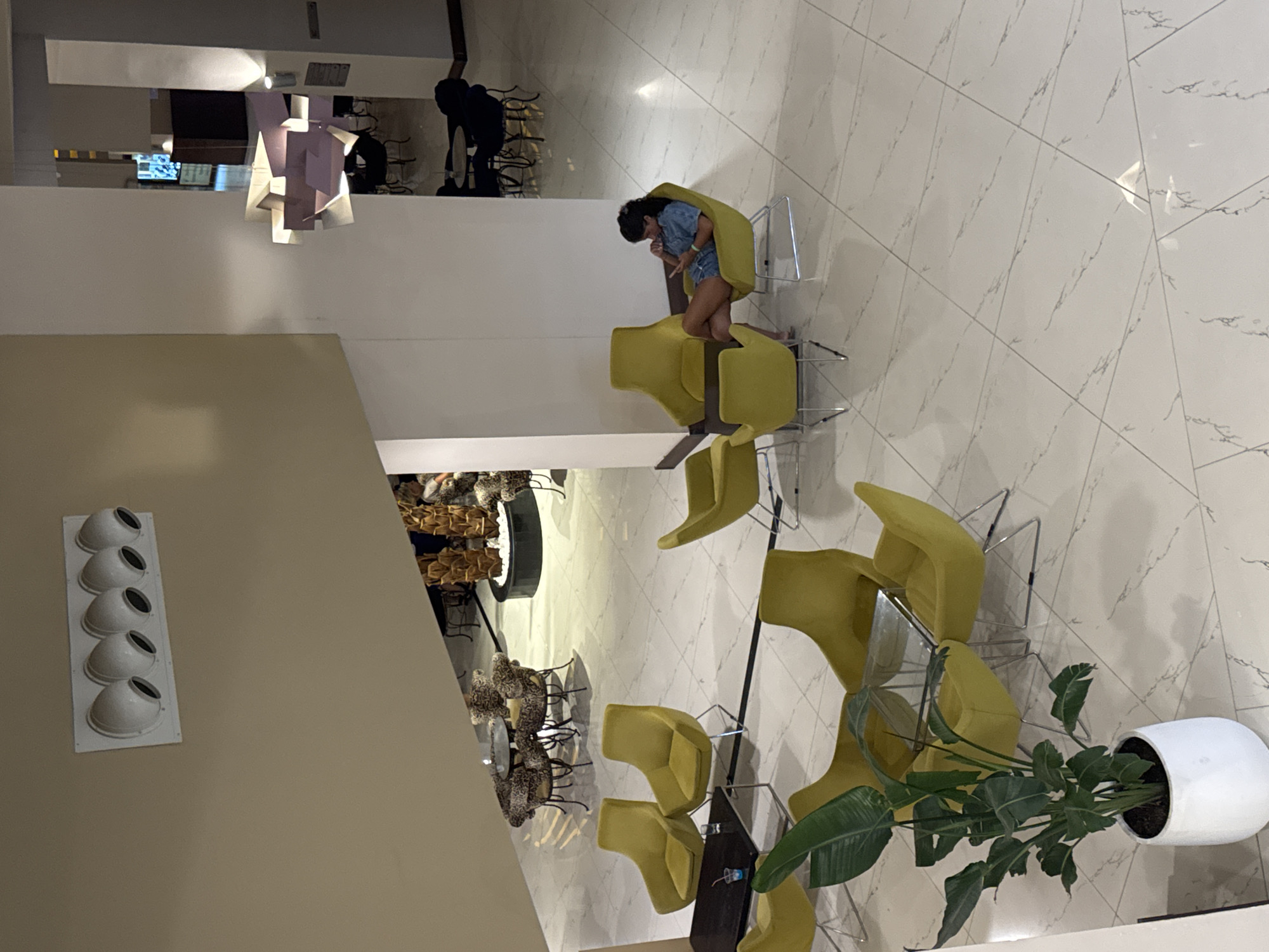 A high-angle shot captures a hotel lobby in Aksu, Turkey, featuring a woman seated in a yellow armchair, engrossed in her phone. The lobby is decorated with a modern aesthetic, including a distinctive geometric light fixture suspended from the ceiling and large potted plants. The floor is tiled in a white marble pattern. The woman, with dark hair pulled back, wears a denim outfit and has her legs crossed as she sits comfortably. Scattered throughout the lobby are additional yellow armchairs and small tables. In the background, a seating area with leopard-print upholstered chairs around a circular table is visible. The overall environment suggests a stylish and contemporary hotel lounge.