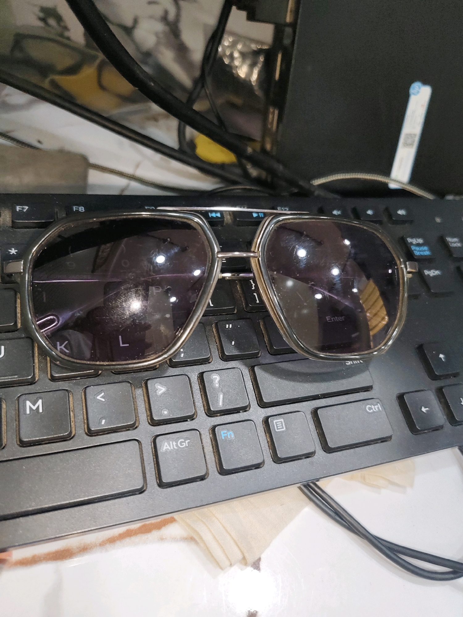 A pair of sunglasses rests on a black keyboard. The sunglasses have a silver frame and dark lenses that reflect the surrounding environment. The keyboard appears to be part of a computer setup, with various keys visible, including "M," "AltGr," and "Enter." Wires are also present, suggesting an active workspace. The background is cluttered with what appears to be office supplies and electronics. The scene is likely indoors, possibly in an office or home environment.