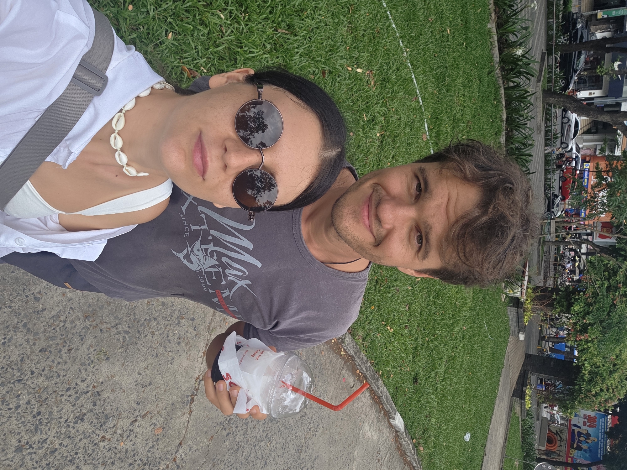 The image is a selfie featuring two individuals: a woman and a man. The woman is wearing sunglasses, a white top, a white shirt with its top buttons undone, and a seashell necklace. The man is wearing a grey t-shirt. He holds a plastic cup with a red straw in his hand.
The setting appears to be outdoors in Thủ Đức, Vietnam. Visible in the background are green grassy areas and urban elements such as sidewalks, trees, parked motorbikes, cars, and buildings.