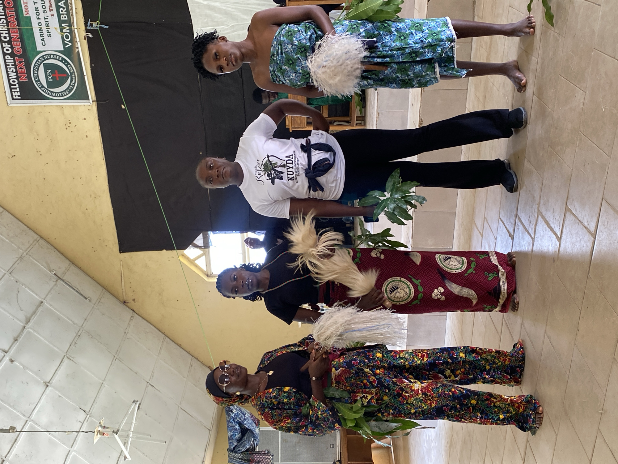 The image shows a group of four women standing indoors, possibly at a church or community hall in Dutsen Kato, Nigeria. The women are of African descent and appear to be participating in a cultural or religious event.
The woman at the front is wearing a colorful, patterned outfit with a headscarf and sunglasses. She holds leafy branches in her hands. Next to her, a woman in a black shirt and a maroon, patterned skirt is also holding leafy branches and a white, fluffy object. The third woman, in a white t-shirt with text and black pants, stands to the left of the maroon-skirted woman. The woman at the back, wearing a blue and green patterned strapless dress, holds a similar white, fluffy object. The overall mood seems festive and ceremonial.
Visible text on a banner in the background reads: "FELLOWSHIP OF CHRISTIAN NURSES NEXT GENERATION", "FCN", "Kufere", "KUYDA", "FUL DEVELOPMENT ASSOC.", "1 Thess. 5:23", and "BEROM WOMEN DEVELOP..."
The indoor setting has tiled floors and walls, and a black backdrop is visible behind the women. Natural light streams in from above, suggesting it is daytime. The presence of leafy branches and traditional attire hints at a local cultural celebration or ceremony.