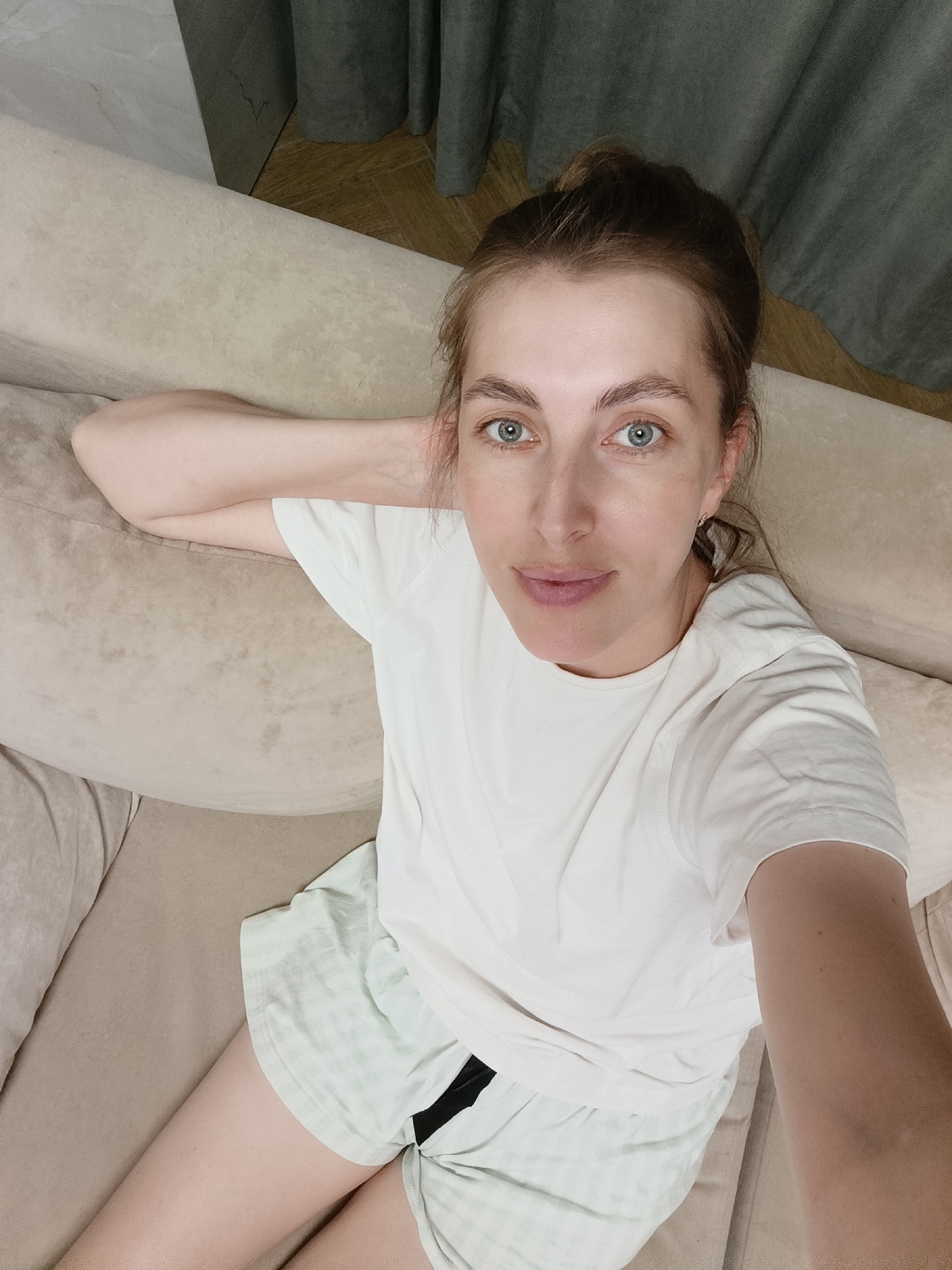 A woman is lying on her side on a couch, holding her head with her arm. She is wearing a white t-shirt and light green shorts. Her hair is pulled back in a bun. The background shows curtains and a wooden floor. The image appears to have been taken in a residential setting, possibly an apartment in Chaikovsky, Russia.