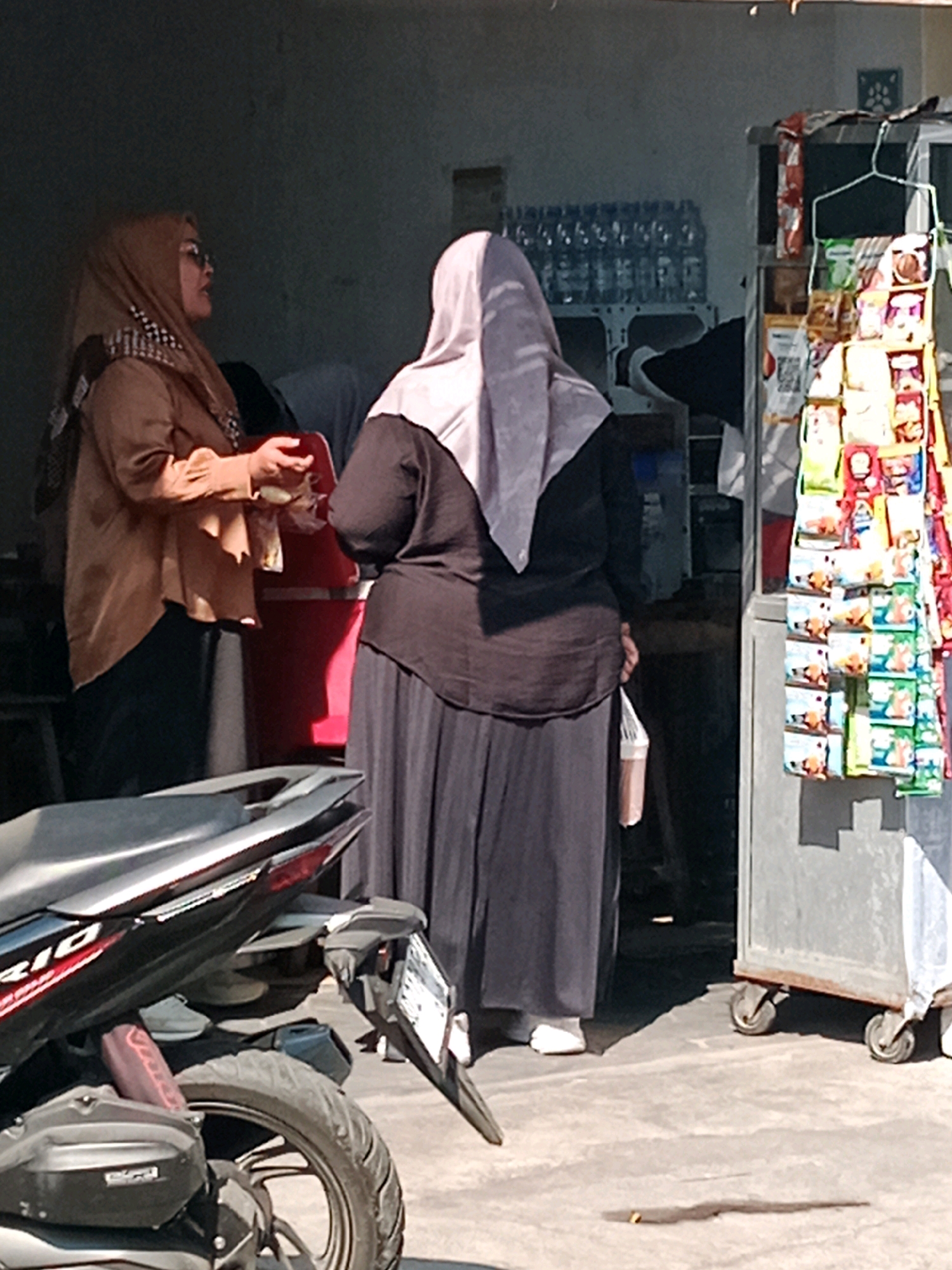 The image depicts an outdoor scene in Cemani, Indonesia, likely near a roadside stall or shop. Two women are the primary subjects. The woman on the left, wearing a light brown hijab, a brown long-sleeved shirt, and dark pants, is holding a red object, possibly a container or bag, and appears to be engaged in a transaction or interaction. She is wearing sunglasses. The woman on the right, wearing a light grey hijab and a dark long-sleeved top with a long dark skirt, is holding a clear plastic bag containing a brown beverage. She is also wearing white slip-on shoes.
In the background, a rack filled with various packaged snacks and drinks is visible, suggesting a small retail establishment. To the left of the women, a red cooler or container is present. A motorcycle, predominantly black with red accents and a license plate, is positioned in the foreground, partially obscuring the lower portion of the scene. The ground appears to be a concrete surface. The overall lighting suggests a sunny day.