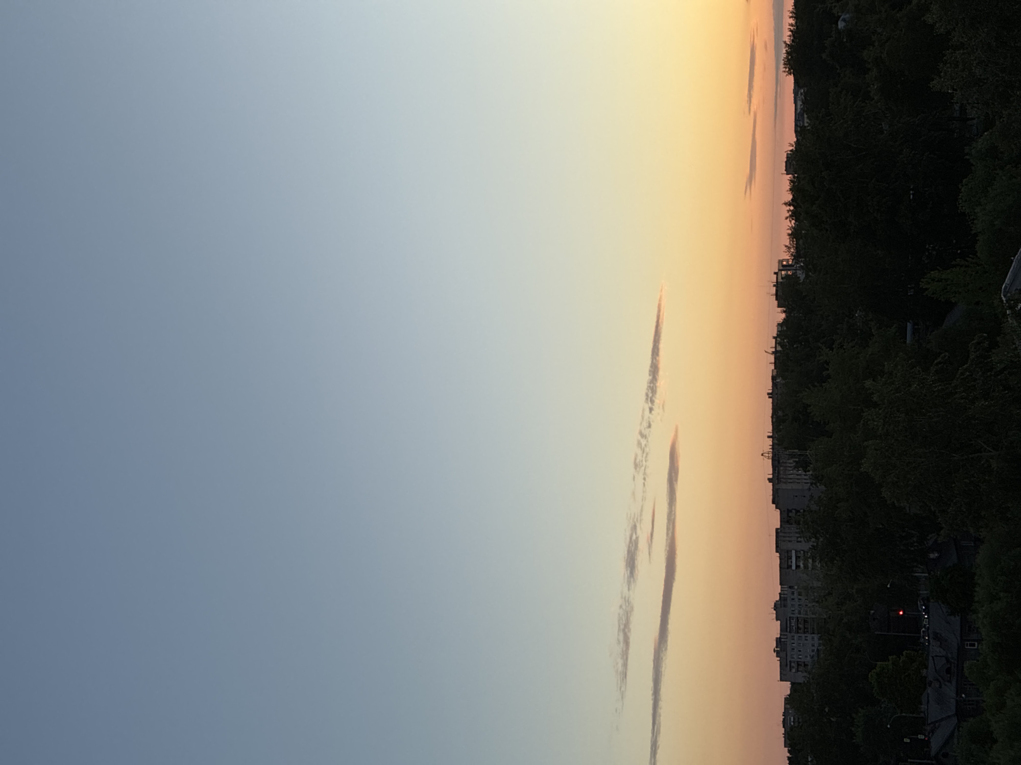 A horizontal, high-angle, wide shot captures a city skyline at dusk. The sky is a gradient of clear blue at the top, transitioning to soft oranges and yellows near the horizon. A few elongated, wispy clouds are present. In the midground, a dense line of dark green trees obscures much of the lower portion of the scene. Above the trees, a series of monotonous, gray apartment buildings are visible, their shapes softened by the dimming light. A faint hint of red from a traffic light can be seen amidst the trees and buildings. The overall impression is one of urban sprawl at the close of day.
