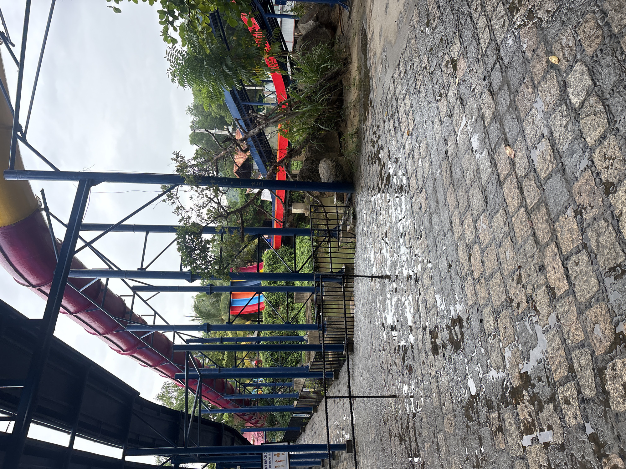 A red and yellow water slide snakes through an outdoor amusement park. The slide is supported by a blue metal frame and is surrounded by lush green trees and foliage. The ground in front of the slide is paved with stone tiles and is wet, with several puddles reflecting the overcast sky. The scene appears to be located in an area with a tropical climate, possibly in Southeast Asia, given the vegetation. The general atmosphere is one of a quiet, possibly rainy day at a water park.