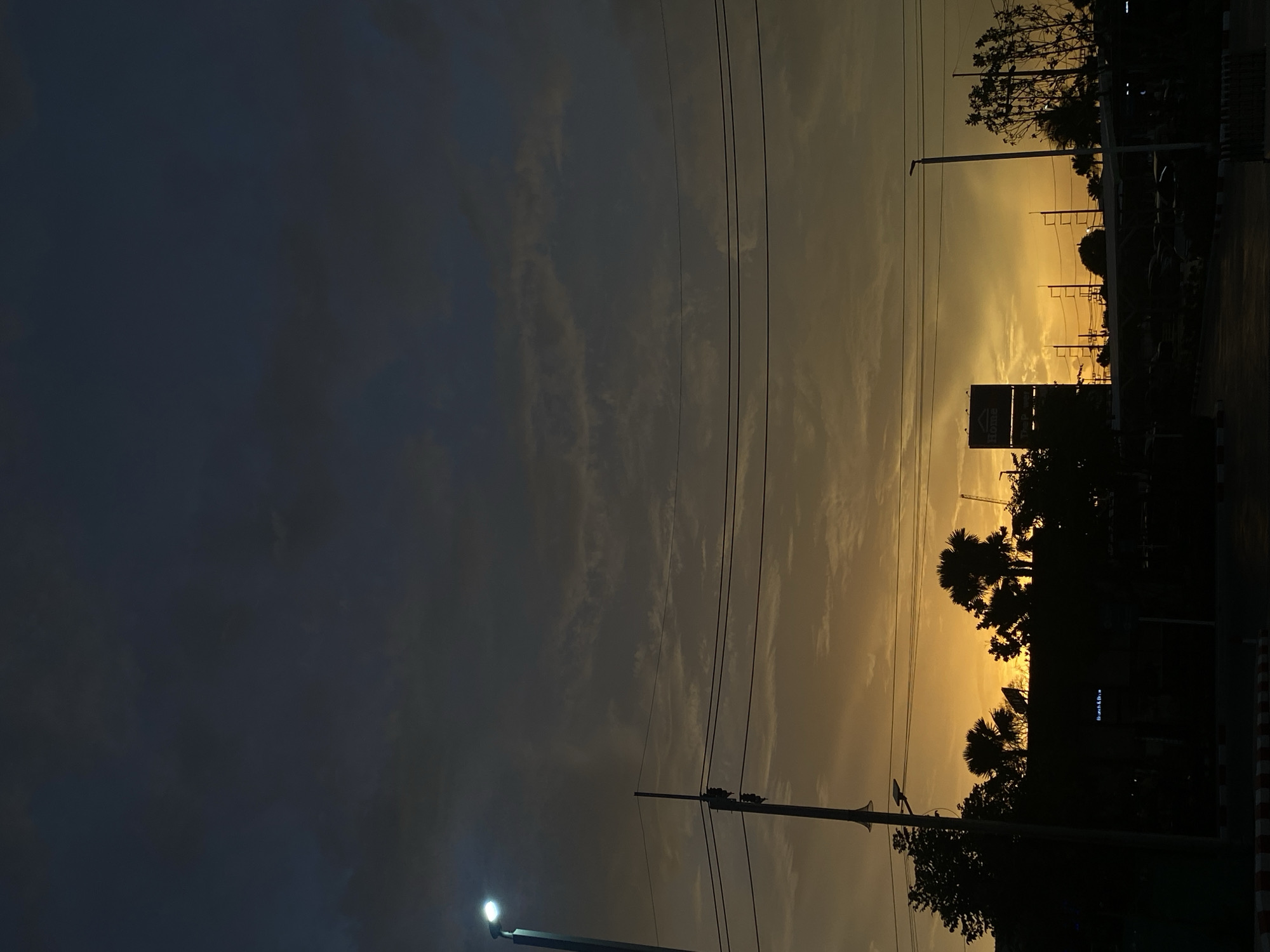 The image shows a sunset scene in Choeng Thale, Thailand. The sky transitions from dark blue-grey on the left to a bright yellow-orange on the right where the sun is setting. Silhouetted against the bright sky are utility poles, power lines, trees, and a building with a billboard. A street light is visible on the lower left side. The scene appears to be near a road, indicated by the edge of the road on the right side.
