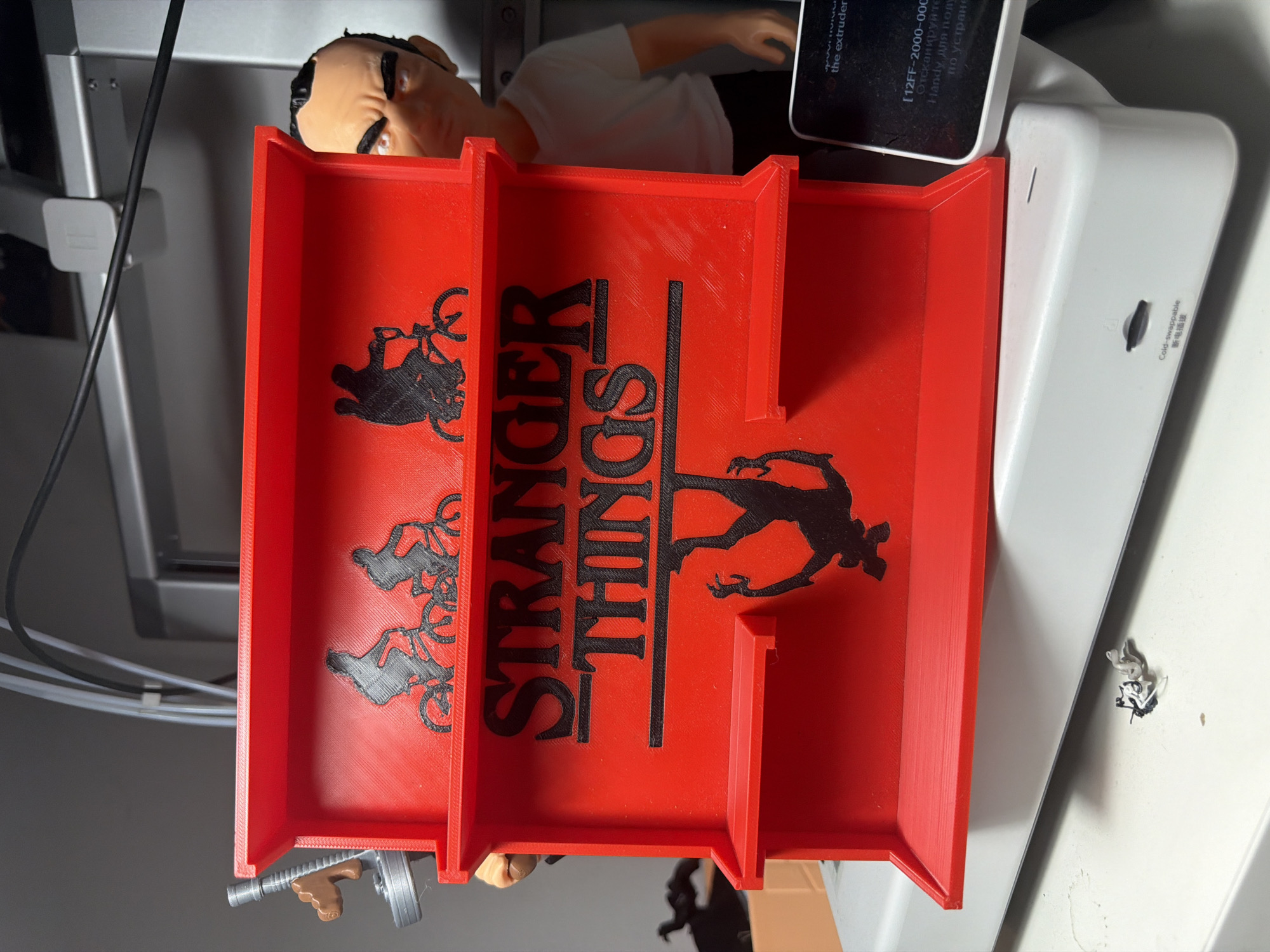 A bright red tray with "STRANGER THINGS" in bold black letters is displayed. The tray is divided into sections, and each section features a black silhouette. One section shows a silhouette of a person on a bicycle, another has a silhouette of the Demogorgon from Stranger Things, and two sections have silhouettes of people on bicycles. A small toy gun is resting on the edge of the tray. In the background, a doll-like figure with tan skin, dark hair, and black eyebrows is visible, wearing a white shirt. To the right of the tray, a white electronic device is partially visible with some text, including "Cold-swappable" and Chinese characters, and other text in a foreign language. The scene appears to be indoors, possibly in a workshop or gaming area, given the presence of electronic equipment and the overall aesthetic. The image is taken from a top-down perspective, with the tray angled towards the viewer.