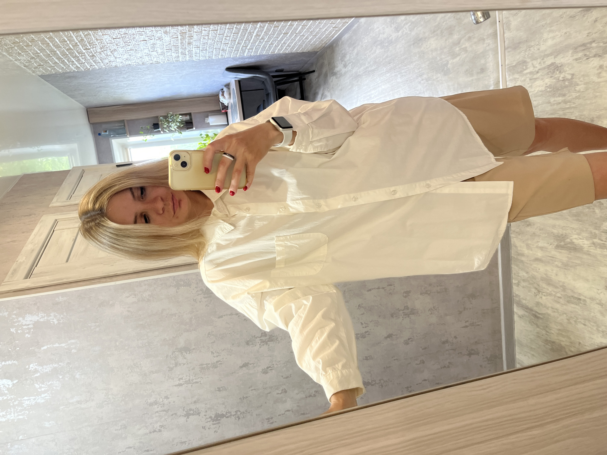 A woman with blonde hair is taking a mirror selfie in what appears to be an apartment hallway. She is wearing an oversized white button-down shirt and tan shorts. She is holding a yellow smartphone in her right hand. In the background, a wooden door is visible on the left, and the mirror reflects a kitchen area with shelves and a chair. The walls have textured wallpaper, and the floor is tiled. The overall scene is dimly lit. There is no visual information in the image to suggest this location is in the city of Chaikovsky, Russia.