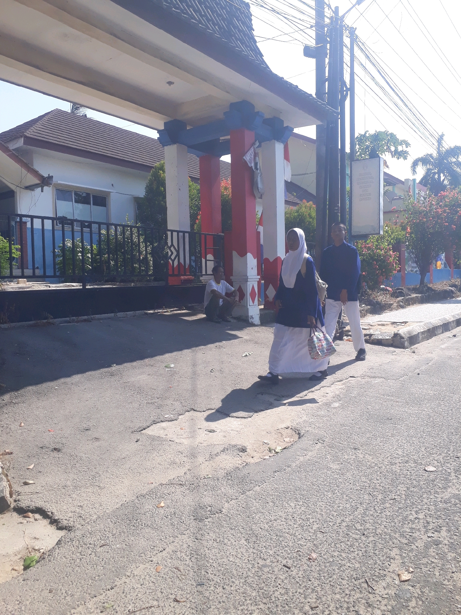 In an outdoor scene on a sunny day, two individuals are walking along an asphalt road. The person on the left is wearing a blue hijab and a white and blue outfit, carrying a patterned bag. The person on the right is wearing a blue shirt and white pants. In the background, there is a building with a tiled roof, a fence, and lush green foliage. A white sign is mounted on a utility pole. Another person is sitting on the ground near the fence. The road is in poor condition, with visible potholes. The scene suggests a pedestrian interaction in a semi-urban or suburban environment.