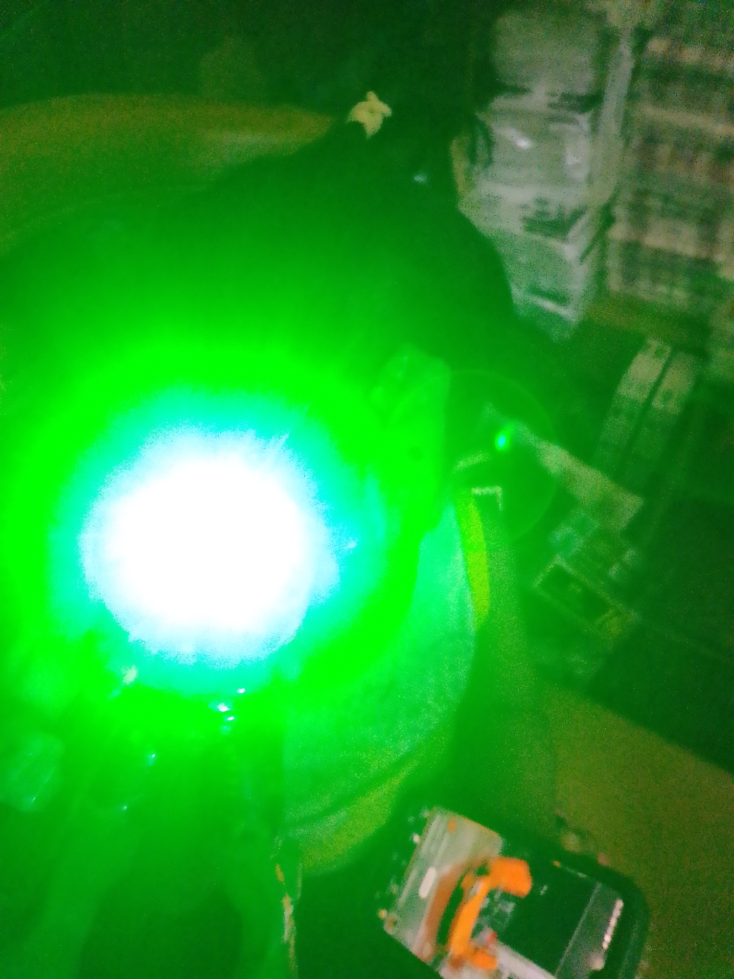 A person is holding a bright green laser pointer, creating a large, intense green glow with a bright white center. The laser beam is directed upwards and outwards. In the background, there appear to be stacks of boxes or similar items. To the right, a smartphone is visible, with some orange components on its screen. The scene is dark, with the dominant illumination coming from the laser. The location is not discernible from the image itself, and therefore cannot be specified as being in Palembang, Indonesia, or any other location.