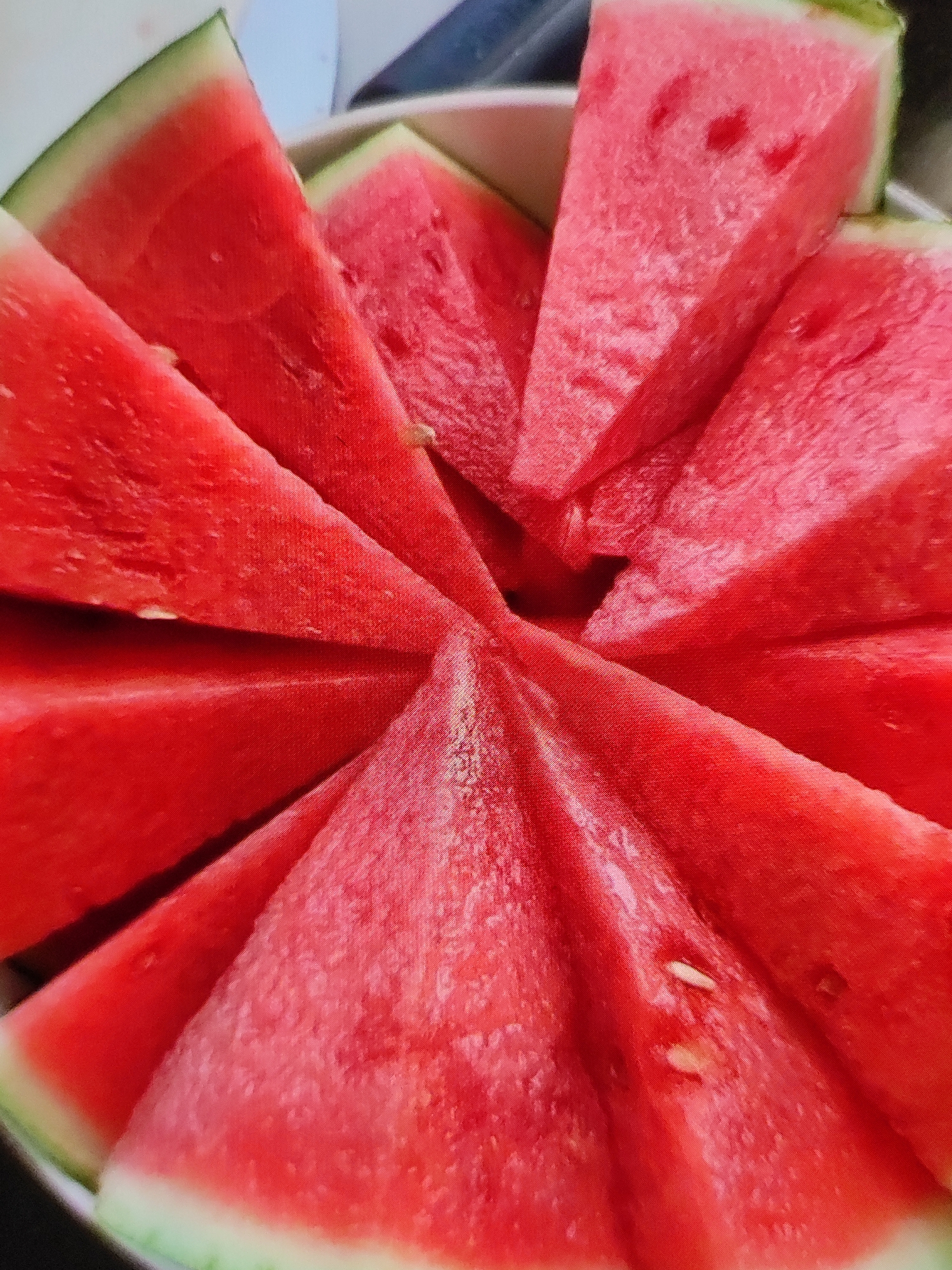 The image shows a close-up view of sliced watermelon arranged in a circular pattern. The watermelon slices are triangular in shape, with bright red flesh and a thin green rind. Some slices have visible seeds. The arrangement suggests the watermelon is served in a bowl or on a platter. The lighting is bright and natural, highlighting the texture and vibrant color of the fruit. There are no people or specific landmarks visible to indicate a location in Tashkent, Uzbekistan. The scene is a still life of fruit.