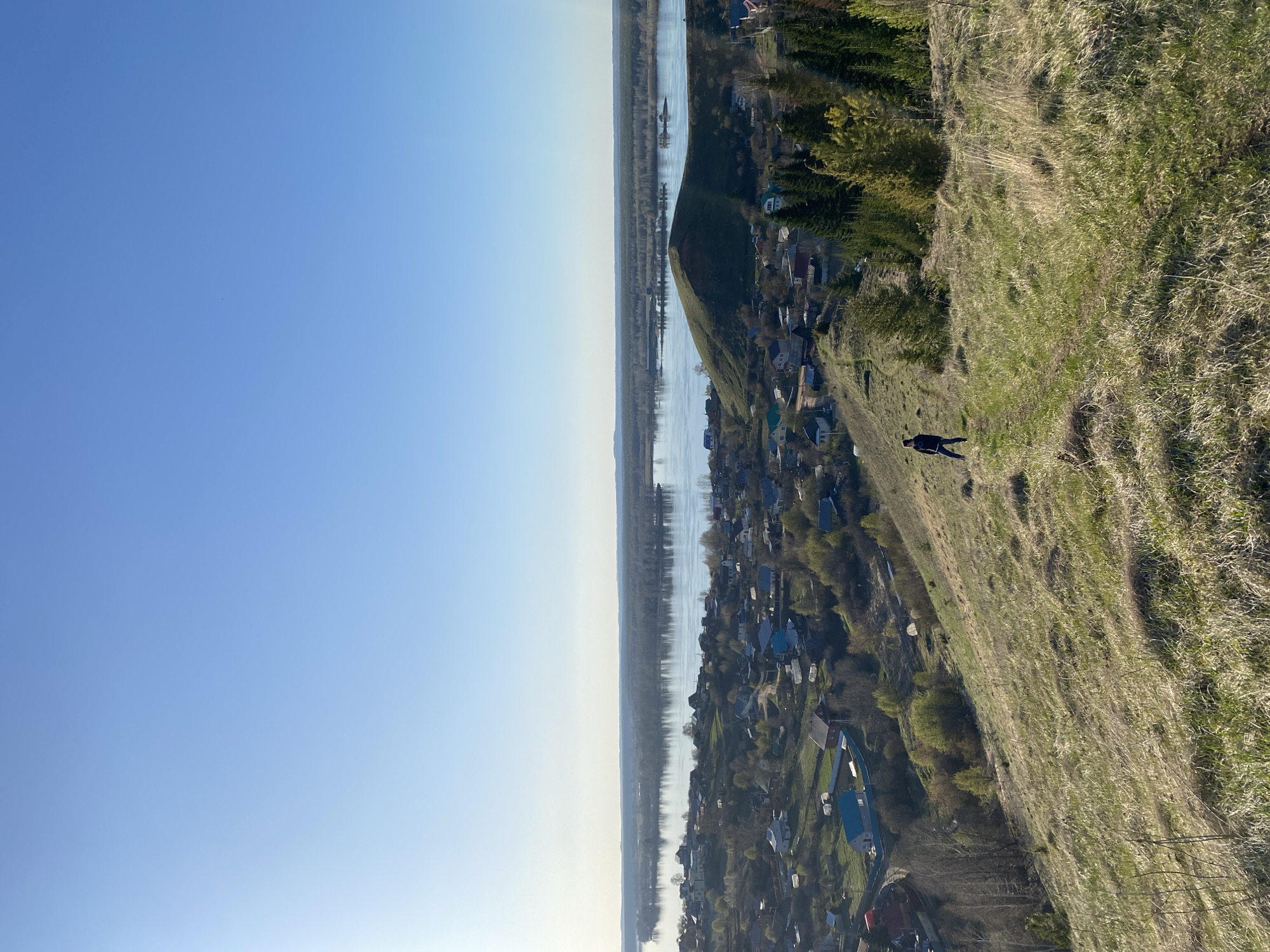 The image presents an elevated view of the village of Olkhovka, Russia, likely captured from a hillside overlooking the town and the adjacent river. A clear blue sky fills the left side of the frame. A wide river stretches horizontally across the middle ground, reflecting the sky and separating the village from the opposite bank. The village is densely populated with houses and buildings, many of which have blue rooftops. The hillside in the foreground is covered with grass and vegetation. A single figure is walking along the hillside.