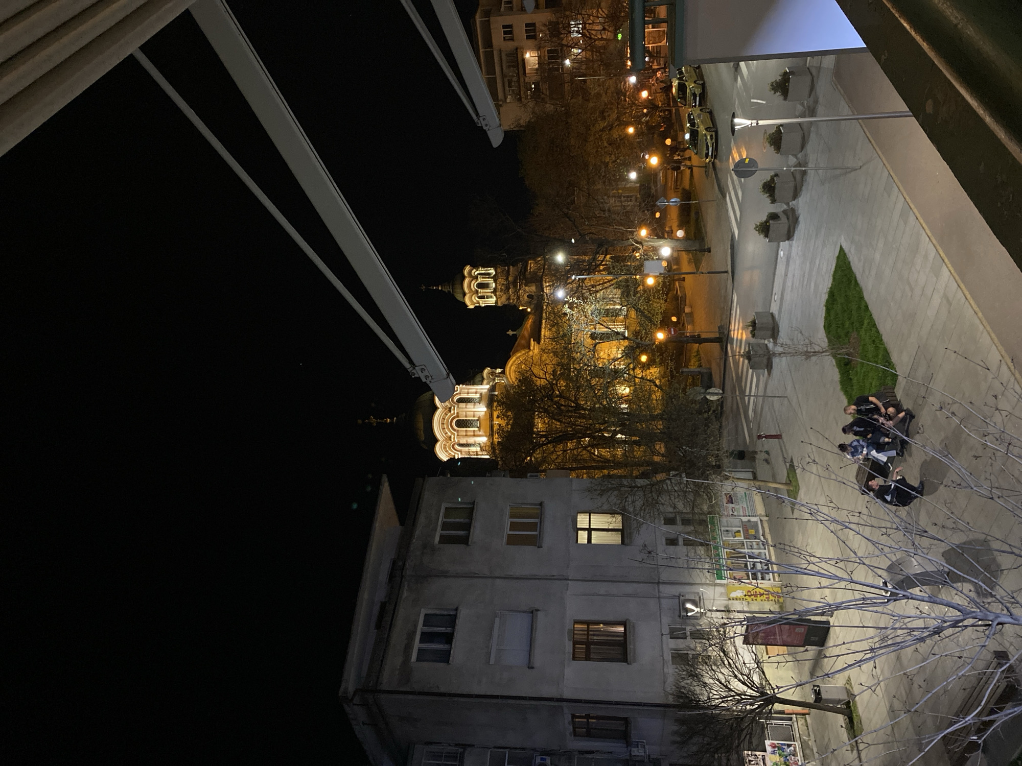View from a window overlooking an illuminated cityscape at night. Prominent is the dome of a church, brightly lit, with a building directly in front of it. A street with parked cars and streetlights runs parallel to the building. A sidewalk has a group of people seated on it near a tree in the foreground. Stara Zagora, Bulgaria.