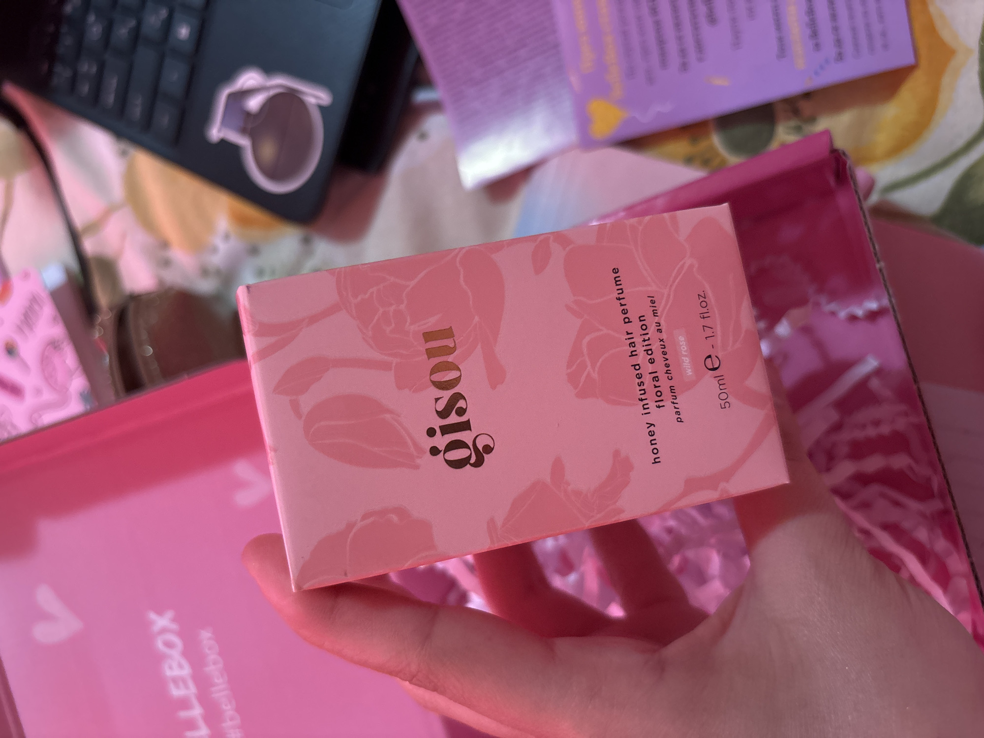 A photograph depicts a hand holding a pink Gisou honey-infused hair perfume box with floral details. The box rests inside a pink "ELLEBOX" branded box. A laptop with a sticker on it is visible in the upper left, and part of a purple box is visible in the upper right. The background includes a floral patterned surface.
