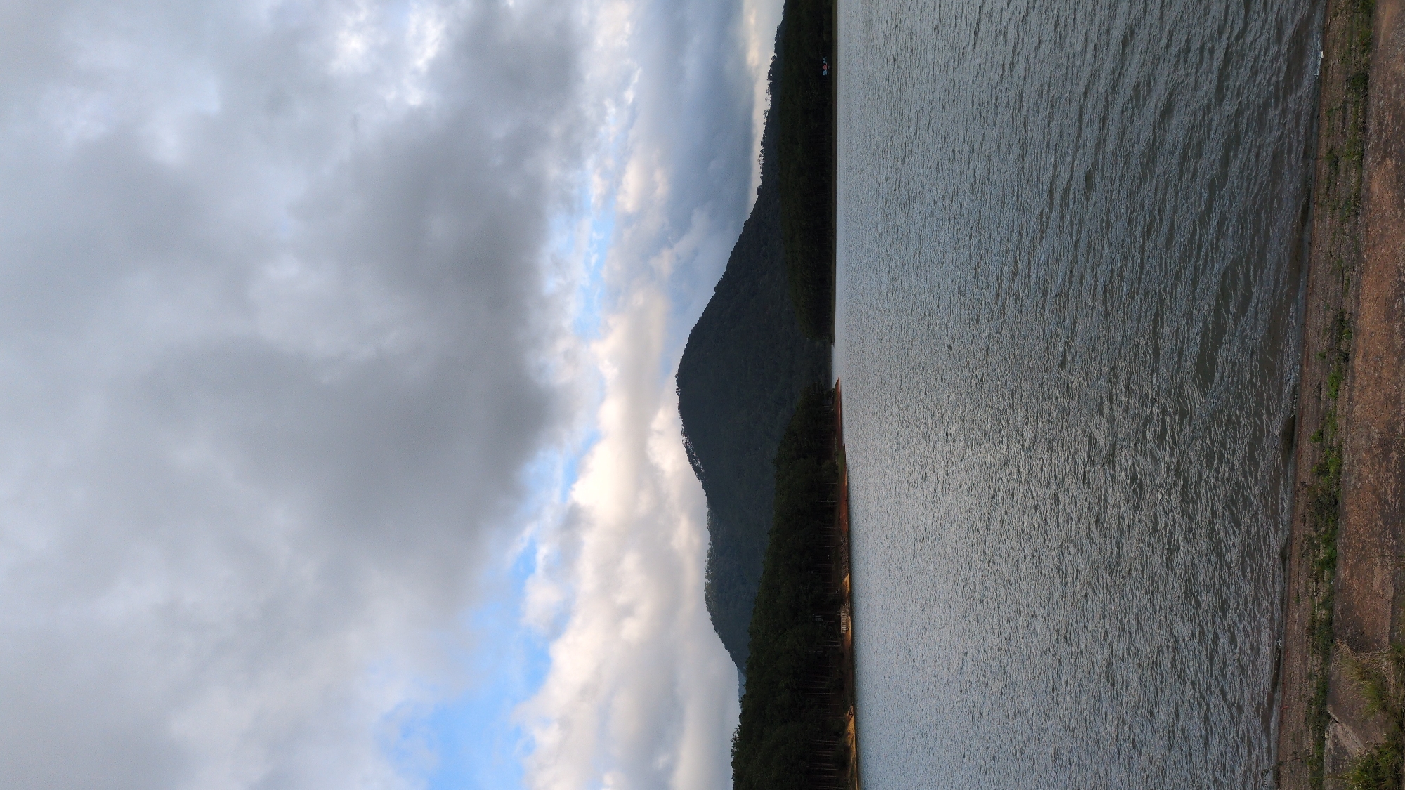 The image captures a wide-angle view of Tuyen Lam Lake in Da Lat, Vietnam. The foreground consists of a textured, reddish-brown concrete embankment with sparse green vegetation. The main body of the image is dominated by the calm, dark blue water of the lake, with subtle ripples on its surface. In the mid-ground, a dense line of dark green pine trees borders the lake, creating a distinct line between the water and the hills beyond. Behind the treeline, the landscape rises into dark green, forested hills, with a prominent, rounded mountain peak dominating the center of the frame. The sky above is filled with dramatic, heavy grey clouds, with patches of lighter grey and occasional glimpses of pale blue sky peeking through. The overall impression is a serene, natural landscape under a cloudy, potentially overcast sky.