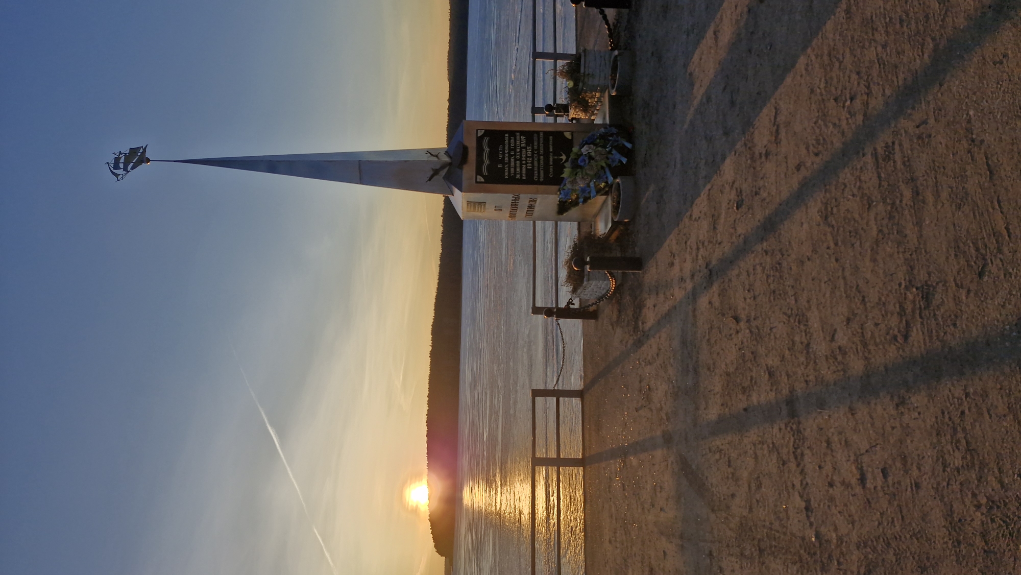Here is a detailed description of the image:
The image shows a monument in Otchor, Russia, at sunset. The monument is a tall, pointed structure topped with a ship weathervane. A plaque is attached to the base of the monument. It is adorned with floral wreaths. In front of the monument, a chain-link fence borders the edge of a body of water, likely a lake or river, reflecting the setting sun. The ground near the monument is bare and sandy, with shadows cast upon it. The sky transitions from blue to orange as the sun sets on the horizon. The plaque inscription translates to "In honor of young volunteers who went to war during the Great Patriotic War in 1942-1945. Death to the fascist invaders."