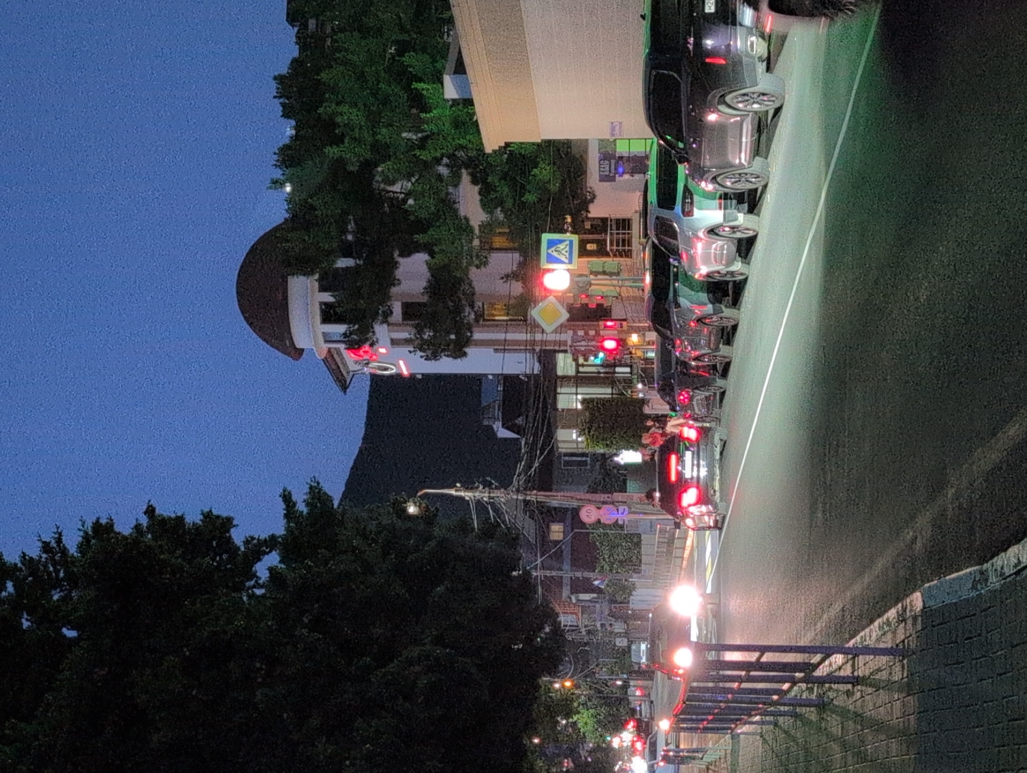 The image shows a street scene at dusk in Gelendzhik, Russia. The street is lined with parked cars and a few pedestrians are visible on the sidewalk. A building with a distinctive domed roof is visible in the background, with a red sign that appears to say "L'One." Traffic lights are visible, indicating an intersection ahead. The sky is a deep blue, and the streetlights and car headlights illuminate the scene. The overall impression is one of a quiet evening in a coastal town.