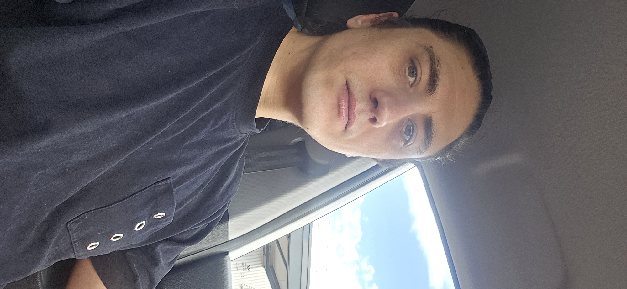 A close-up, medium shot captures a person from the chest up, seated in the passenger seat of a car. The individual, with dark hair pulled back, is looking directly at the camera. They are wearing a dark blue t-shirt with a pocket adorned with three silver grommets. The background shows a bright blue sky with scattered white clouds, suggesting an outdoor setting. Part of the car's interior, including the window frame and seatbelt, is visible. The location is identified as Bous, Germany.
