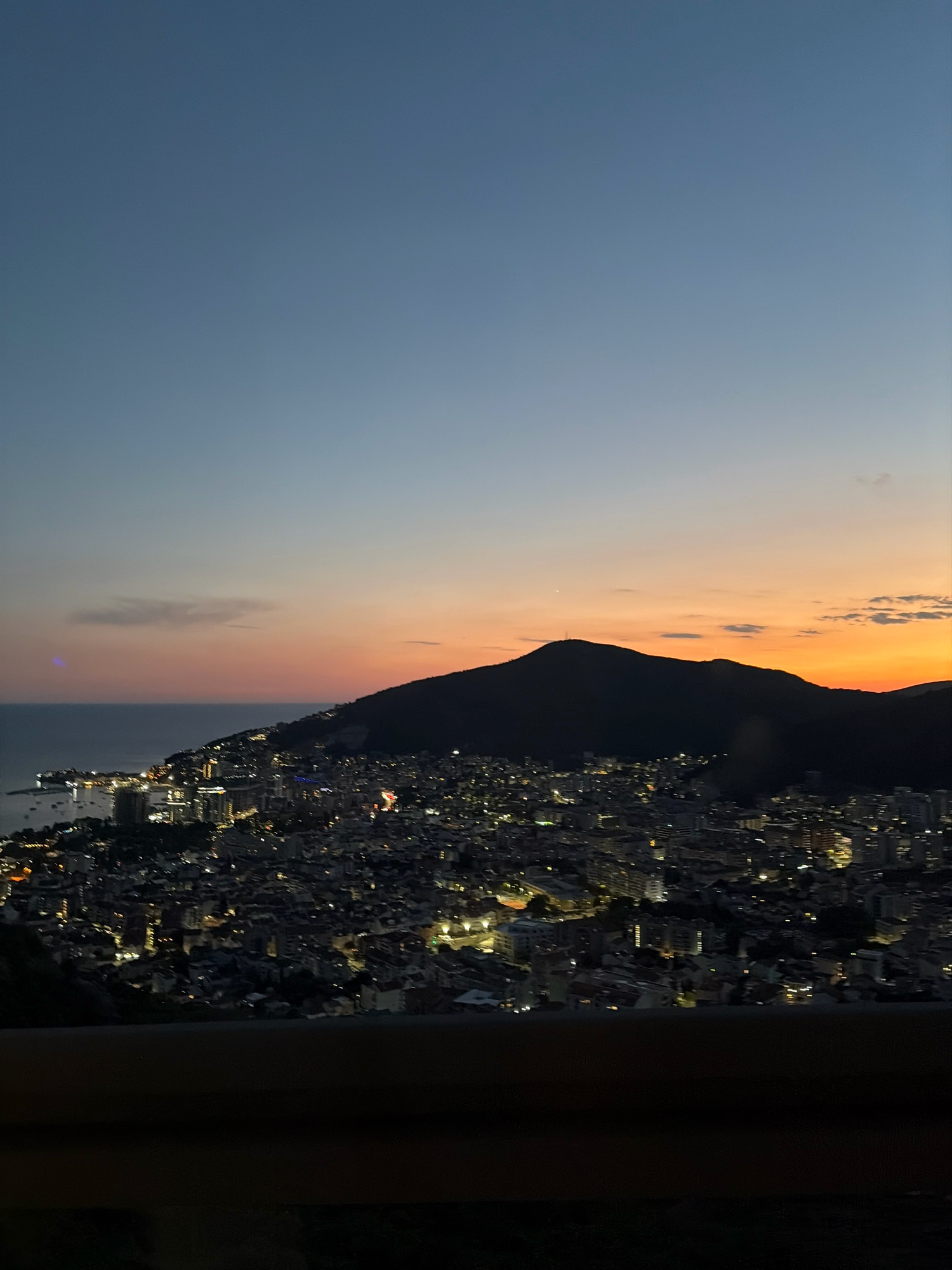 The image is a nighttime aerial view of the city of Budva, Montenegro, during twilight. The sky transitions from a deep blue at the top to orange and pink hues near the horizon, indicating sunset or sunrise. A dark, silhouetted mountain range dominates the right side of the frame, with its peak visible against the colorful sky. Below the mountain, the city of Budva is illuminated by thousands of lights, showcasing the urban sprawl. Buildings of various sizes and shapes are densely packed together, creating a twinkling cityscape. The coastline is visible on the left, with the dark water of the Adriatic Sea meeting the shore. A few boats are anchored in the harbor, their lights reflecting on the water. In the foreground, a dark, blurry structure, likely a railing or barrier, obscures the bottom portion of the image. There are no people visible in the scene. The overall impression is one of a vibrant coastal city coming alive at night under a dramatic sky.