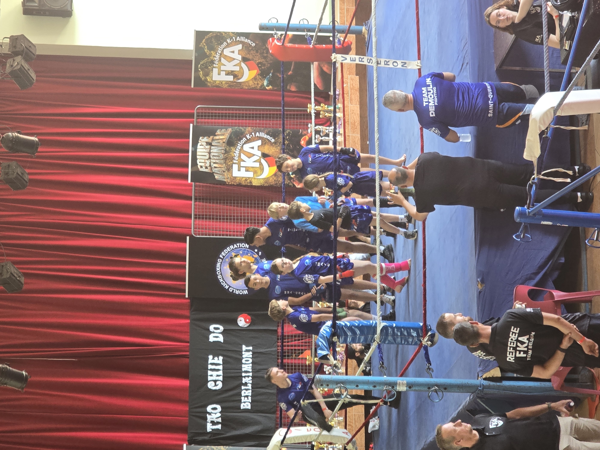The image depicts a boxing ring in Berlaimont, France, with participants, referees, and spectators. A red curtain hangs behind the ring. Banners with "THO CHIE DO BERLAIMONT," "WORLD KICKBOXING FEDERATION," and "EQUIPE NATIONALE FKA Fédération K-1 Alliance" are visible. Individuals in matching blue uniforms stand near the ring. One man wears a shirt with "TEAM DEMOULIN FIGHTING SAINT-QUENT" on the back, while another wears a "REFEREE FKA TEAM BELGIUM" shirt.