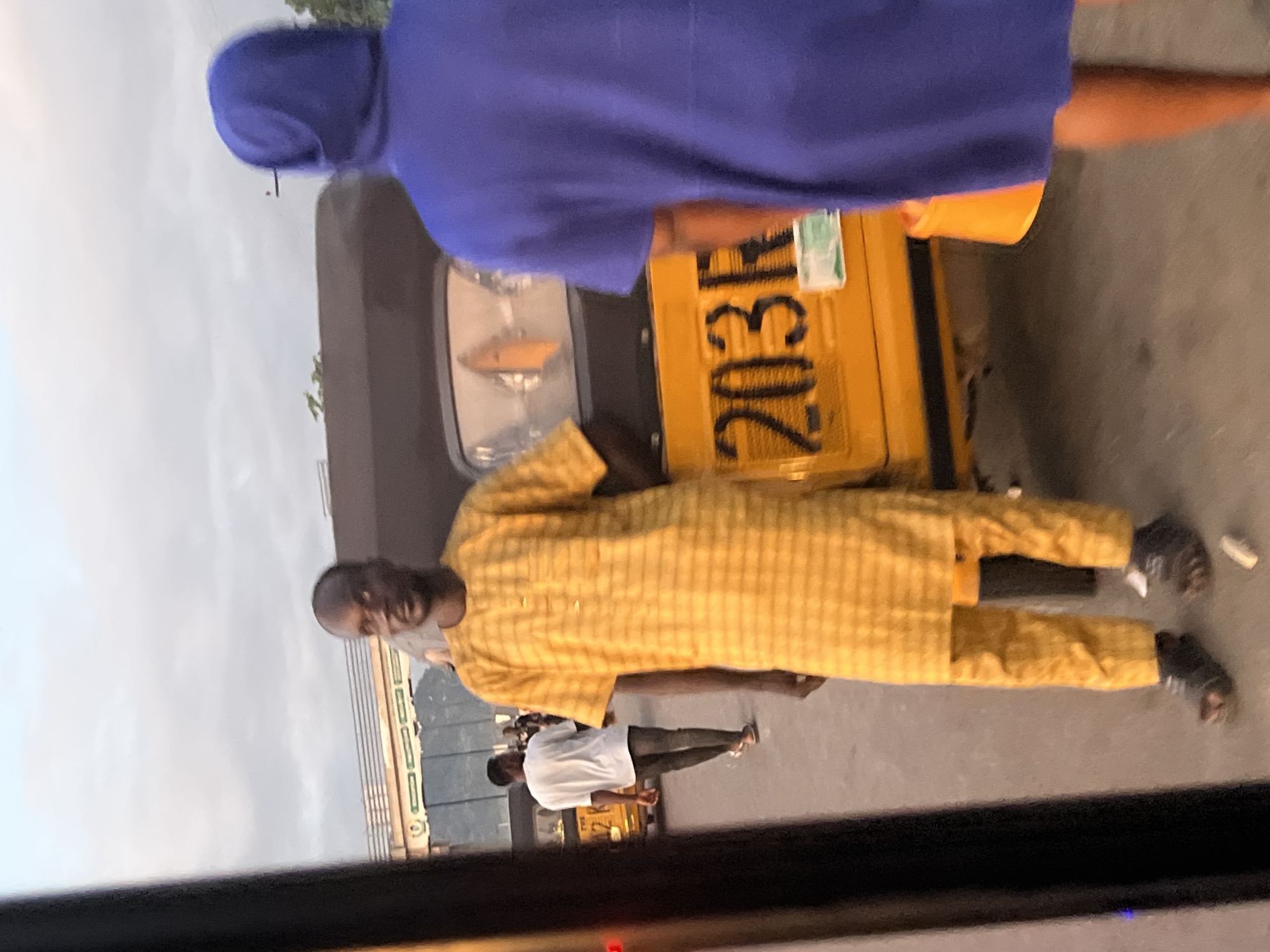 The image depicts an outdoor urban scene in Howayi, Nigeria, under an overcast sky, suggesting either dawn or dusk.
In the mid-foreground, a dark-skinned adult male stands, facing slightly right and looking towards the camera. He is dressed in a yellow traditional outfit featuring a checkered pattern, consisting of a long-sleeved tunic and matching trousers. He wears dark sandals. To his right, partially obscuring a yellow three-wheeled vehicle, an individual stands with their back mostly to the camera, wearing a loose-fitting, royal blue hooded garment that covers their head and body.
Behind the yellow-attired male and to the left, another dark-skinned adult male is walking away from the camera, towards the left side of the frame. He is wearing a white short-sleeved t-shirt, dark trousers (possibly jeans), and light-colored shoes.
The primary vehicle in the scene is a yellow three-wheeled auto-rickshaw, commonly known as a Keke Napep. Its rear is visible, featuring a dark brown or black roof section and a yellow lower body. Black numbers "203R" are prominently displayed on a yellow panel at the rear, consistent with a license plate area. A small green and white paper document is affixed to the right of these numbers. The left taillight shows an illuminated orange indicator.
The ground surface appears to be paved or asphalt. In the background, a bridge or overpass structure with a railing is visible, featuring green signage with white text and reflective panels underneath. Some indistinct green foliage is present near the bridge. A thick, dark vertical structure partially frames the scene on the far left.