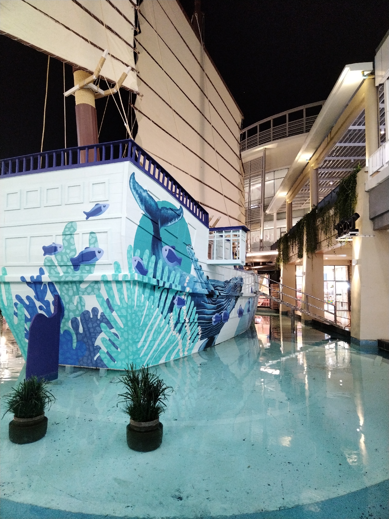 The image shows a decorative Chinese junk boat replica located in a shallow pool of water. The boat is painted with a marine mural featuring a whale's tail and coral. Two potted plants are visible in the foreground, situated in the water. Behind the boat, there is a modern building with multiple levels, balconies, and illuminated windows. The scene is likely an outdoor area or a covered atrium within a commercial or entertainment complex, possibly in a tropical location given the style of the building and the tropical plants. The setting appears to be Patong, Thailand, a popular tourist destination known for its vibrant nightlife and commercial establishments.