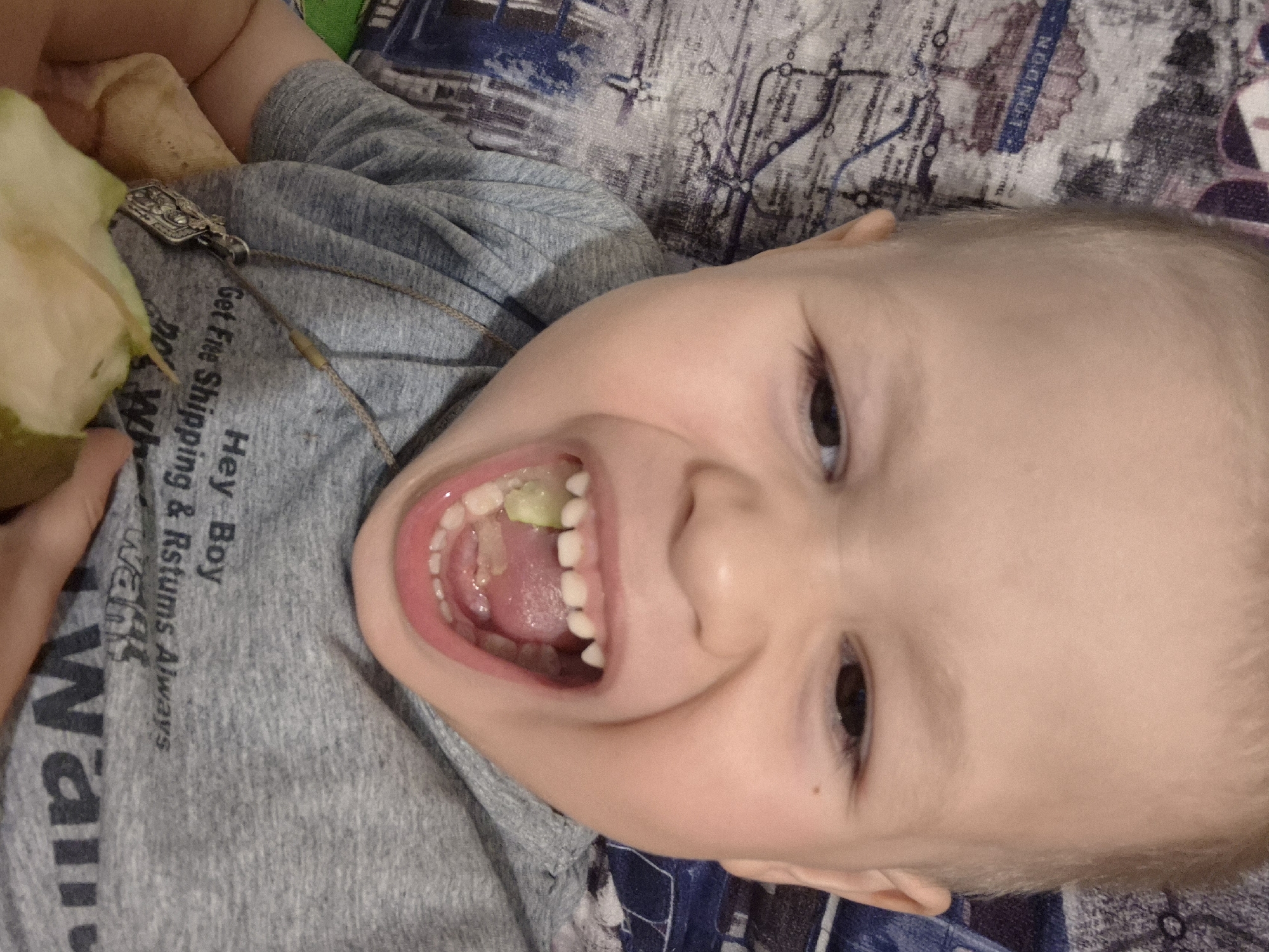 A close-up, high-angle shot captures a joyful young child, likely a toddler, lying down and looking directly at the camera with a wide, open-mouthed smile. The child has fair skin, light-colored hair, and dark eyes. A piece of green fruit, possibly a pear, is visible on their tongue, and a larger portion is held in their left hand. The child is wearing a grey t-shirt with text that reads "Hey Boy", "Get Free Shipping", "Returns Always", and partially "Wail". A silver-colored, ornate bolo tie or pendant necklace rests on their chest. The child is positioned on bedding patterned with what appears to be a map or schematic, featuring the clearly visible word "LONDON" among other indistinct lines and text. The bright, indoor lighting suggests it is daytime, and the child's expression conveys happiness or excitement. The scene is set indoors, likely in a home in Chaikovsky, Russia.