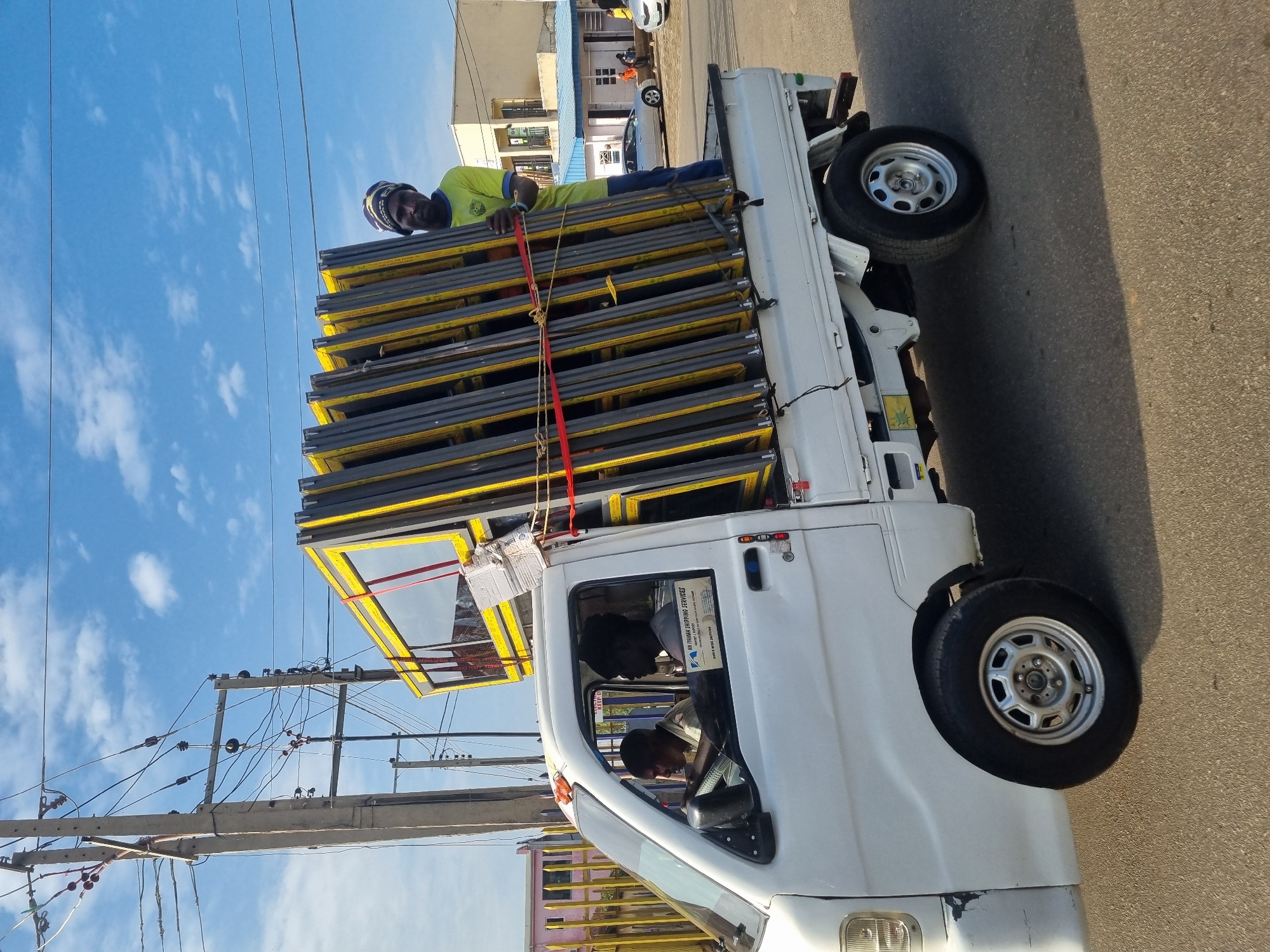 A white truck in Jos, Nigeria, is overloaded with stacked window frames, secured with a red strap, on a city street. A person in a yellow shirt and headgear is perched atop the load. Two people are inside the cab. Power lines and a blue, slightly cloudy sky are visible above.
