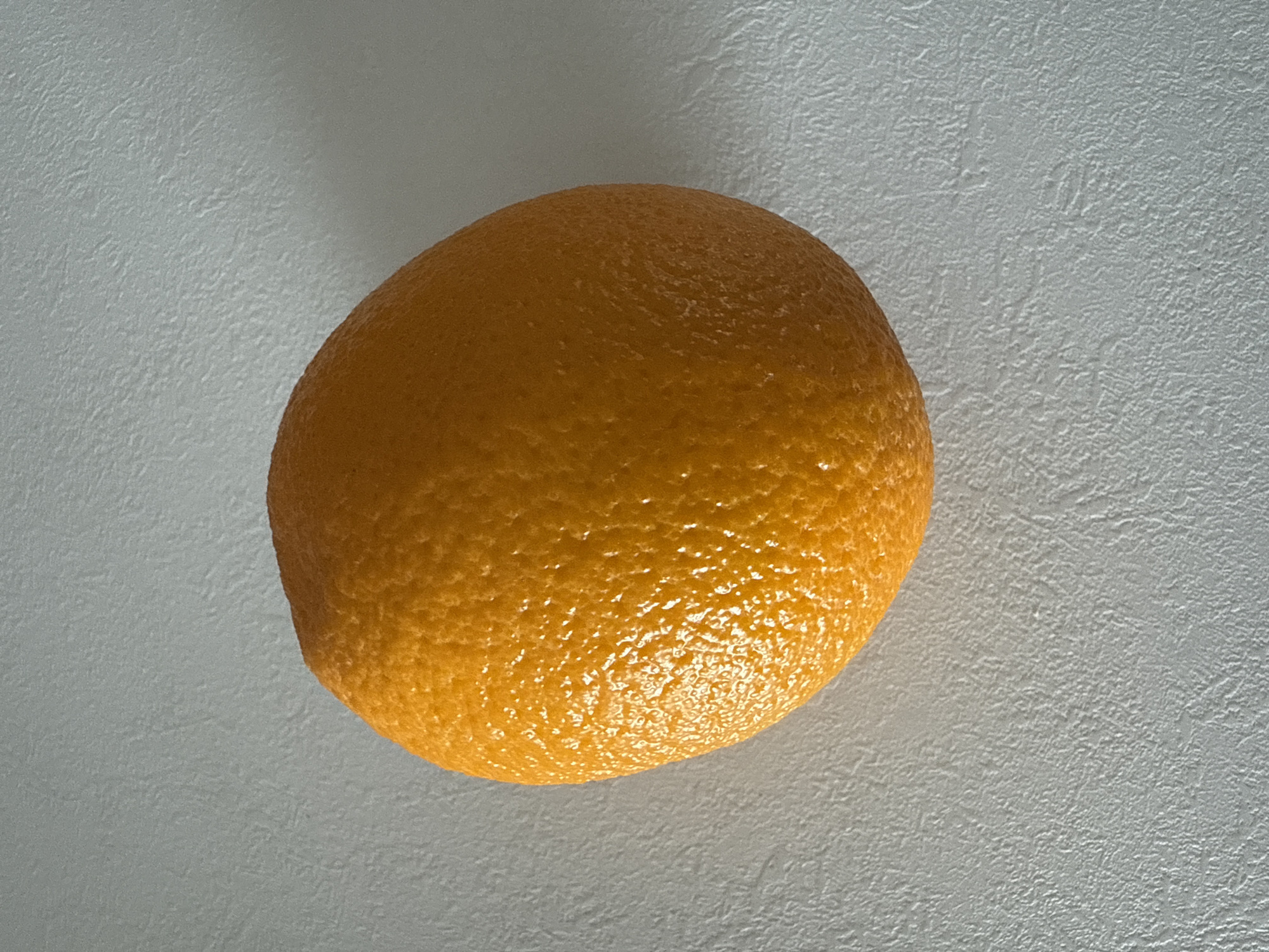 An orange is positioned on a white surface. There is no indication of the location being in Votkinsk, Russia. No people, actions, or interactions are present in the image. The environment consists solely of the white surface and the orange.