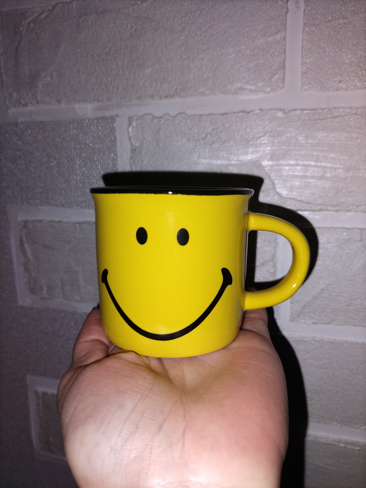 A human hand holds a bright yellow ceramic mug with a black smiley face design against a textured, light-colored wall. The mug features two black circular eyes and a wide, upward-curving black line for a smile, with a black rim around its top edge. A matching yellow handle is visible on the right side of the mug. The hand supports the bottom of the mug, with palm lines and skin texture clearly visible. The background wall has a distinct brick-like pattern with visible mortar lines, suggesting either real bricks or textured wallpaper. A prominent, dark shadow of the mug and hand is cast onto the wall, indicating an artificial light source, making the time of day indeterminate. The scene appears to be indoors. No visible text is present in the image. The smiley face on the mug conveys a cheerful emotion.