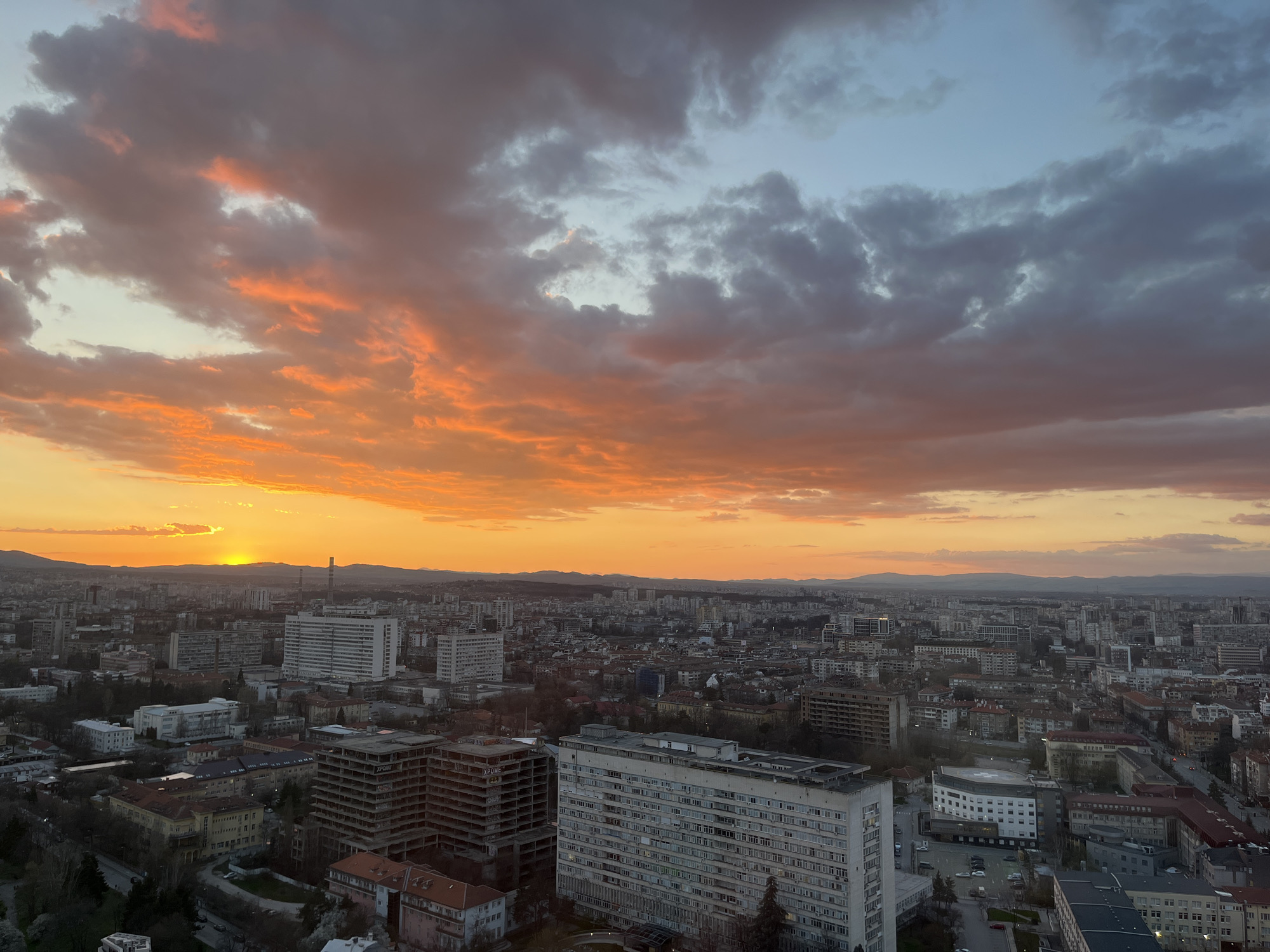 The image captures an aerial view of Sofia, Bulgaria, at sunset. The sky is filled with orange and gray clouds. Below the horizon is a silhouette of mountains. The urban landscape consists of buildings of various sizes and architectural styles. There are many apartment buildings and hospitals present. A major road is visible in the lower right, with moving cars on it. The scene conveys a sense of urban life and the natural beauty of the sunset.