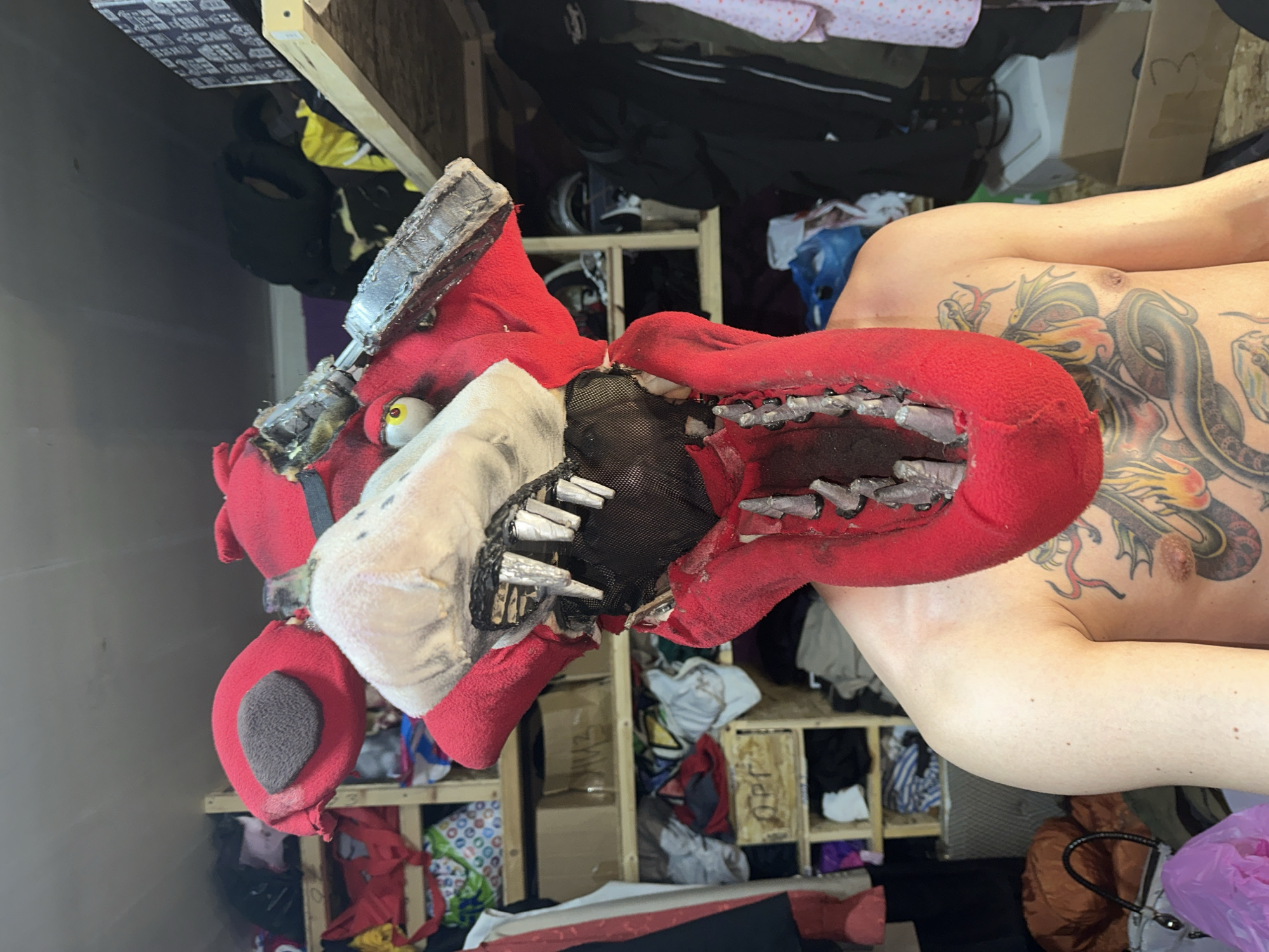 An individual is depicted shirtless, from the mid-torso upwards, wearing a large, elaborate mask in a cluttered indoor setting. The individual exhibits extensive tattoos across the chest and left shoulder, prominently featuring a multi-colored dragon or serpent intertwined with flames.
The mask, covering the entirety of the wearer's head, is predominantly red, fuzzy, and constructed to resemble a worn animatronic fox, consistent with the character Foxy from "Five Nights at Freddy's." It features a dramatically open mouth revealing multiple jagged, metallic teeth on both upper and lower jaws, with a black mesh interior. One yellow eye with a black pupil is visible on the wearer's right side. The other eye socket is covered by a dark, textured patch. A rounded red ear with a dark grey inner surface is visible on the mask's left side. Attached to the mask's right side is a silver-grey, metallic, segmented component, appearing as an exposed or damaged mechanical element. The red fabric of the mask displays signs of wear and discoloration.
The background consists of a densely packed interior room, identified as Saratov, Russia, likely a storage or workshop area. Numerous roughly constructed wooden shelves made from plywood or particle board are visible. These shelves are laden with assorted items including folded and hanging clothing of various colors, cardboard boxes (one labeled "HM3" and a wooden crate marked "OPT"), assorted bags including a white handbag and a pink plastic bag, and other miscellaneous objects. The lighting is internal.