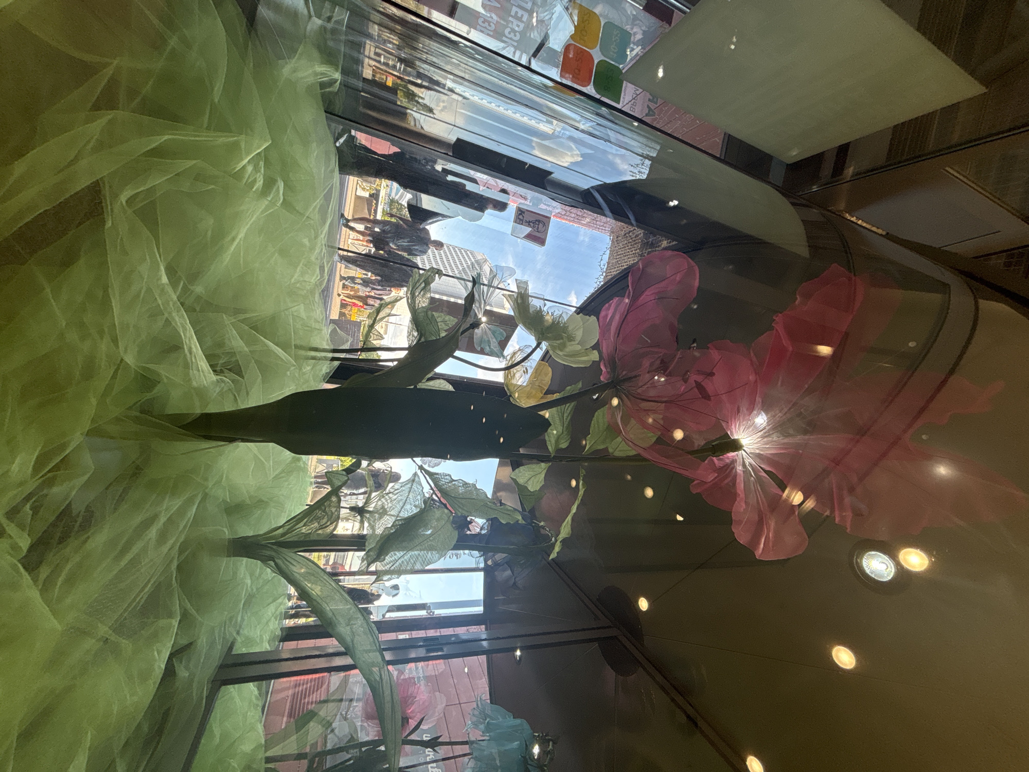 The image captures a vibrant, layered scene, appearing to be viewed through a large glass display or window in an urban setting in Minsk, Belarus. The original image is oriented sideways, with the horizon appearing vertical.
Inside the glass, a prominent artificial floral display features large, stylized flowers crafted from sheer, colorful fabrics in shades of pink, white, and yellow. These are accompanied by green fabric leaves and stems. A significant portion of the left side of the display is filled with draped and bunched light green tulle or netting, adding a soft texture. Several circular spotlights from the interior ceiling or structure reflect brightly off the glass surface, creating points of light.
Through the glass, an exterior urban scene is visible under a clear, bright blue sky with some white clouds, indicating daytime. Indistinct figures of people can be seen walking on a sidewalk, and modern buildings with glass facades rise in the background.
The scene is characterized by complex reflections on the glass, which superimpose interior elements over the exterior view. A notable detail is the clear reflection of a KFC bucket logo. Various fragments of Cyrillic text are visible, both as reflections and potentially on distant signage. These include words like "ТВЕР" (TVER), "МАРК" or "МАРТ," and numerical sequences such as "19-55" and "70-55" displayed within colored squares (green, orange, blue), possibly indicating prices or phone numbers. The abbreviation "BYN" (Belarusian Ruble) is also faintly discernible.