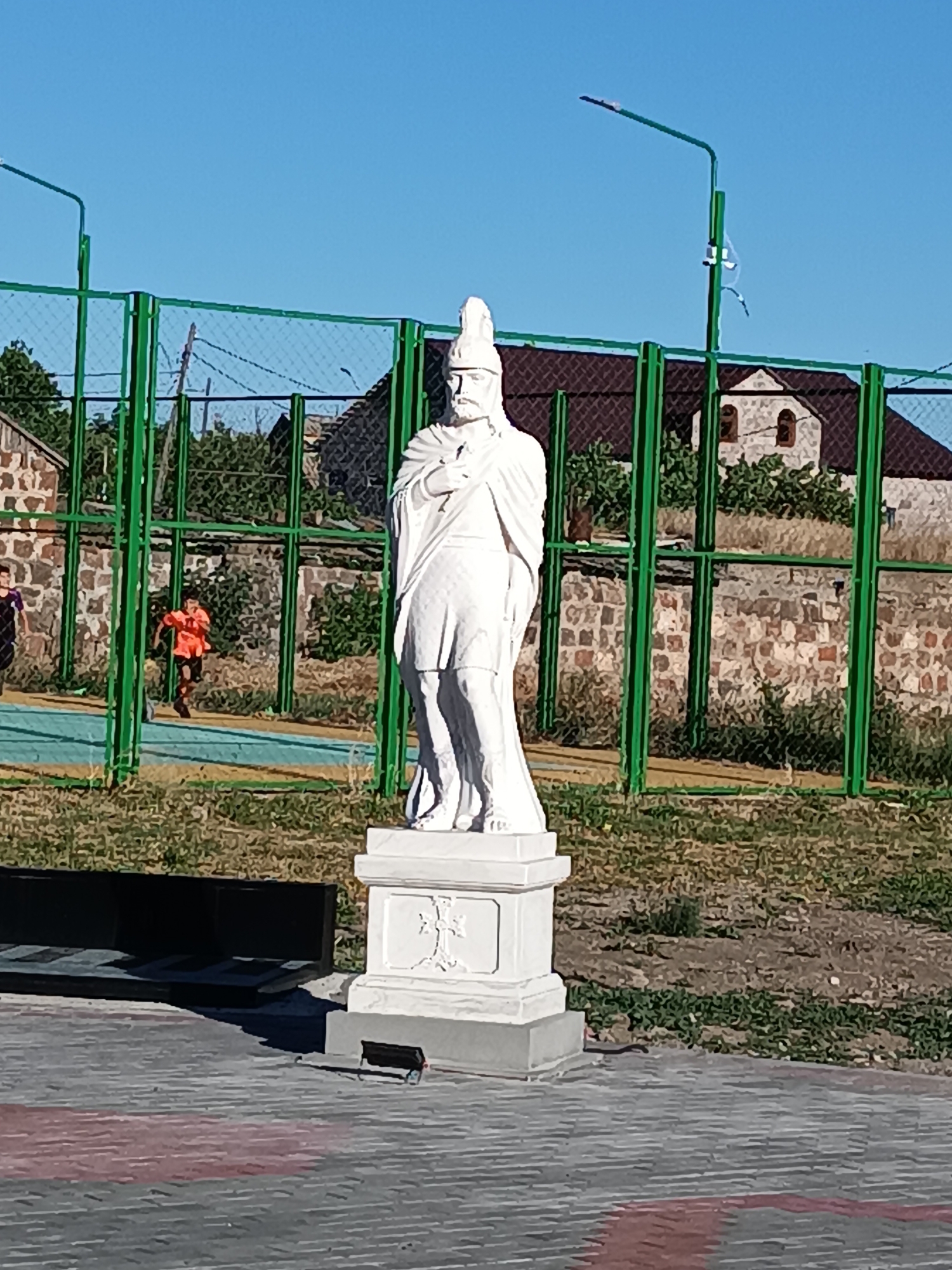 A white stone statue of a historical figure stands prominently in the foreground of the image. The figure is depicted wearing a helmet and a cloak draped over one shoulder, with a hand resting on their chest. The statue is mounted on a multi-tiered pedestal, which features a carved cross on its front.
Behind the statue, a green metal fence encloses an area that appears to be a sports field or court, with a blue-colored surface visible. Two young boys are engaged in activity on this court. One boy, wearing an orange shirt and dark shorts, is running with a ball at his feet, suggesting a game of soccer is in progress. Another child, wearing a dark shirt, is visible to the left of the frame.
Beyond the fence, there are buildings with stone walls and dark roofs, along with trees and vegetation. The sky is a clear, bright blue, indicating a sunny day. The lighting suggests it is daytime, possibly late morning or afternoon. The scene is set outdoors, with paved ground in the immediate foreground, characterized by a patterned surface of gray and red interlocking tiles. A black structure, possibly a bench or planter box, is visible to the left of the statue. The overall atmosphere appears casual and recreational, with the statue serving as a formal centerpiece within a lively outdoor setting. There is no visible text in the image.