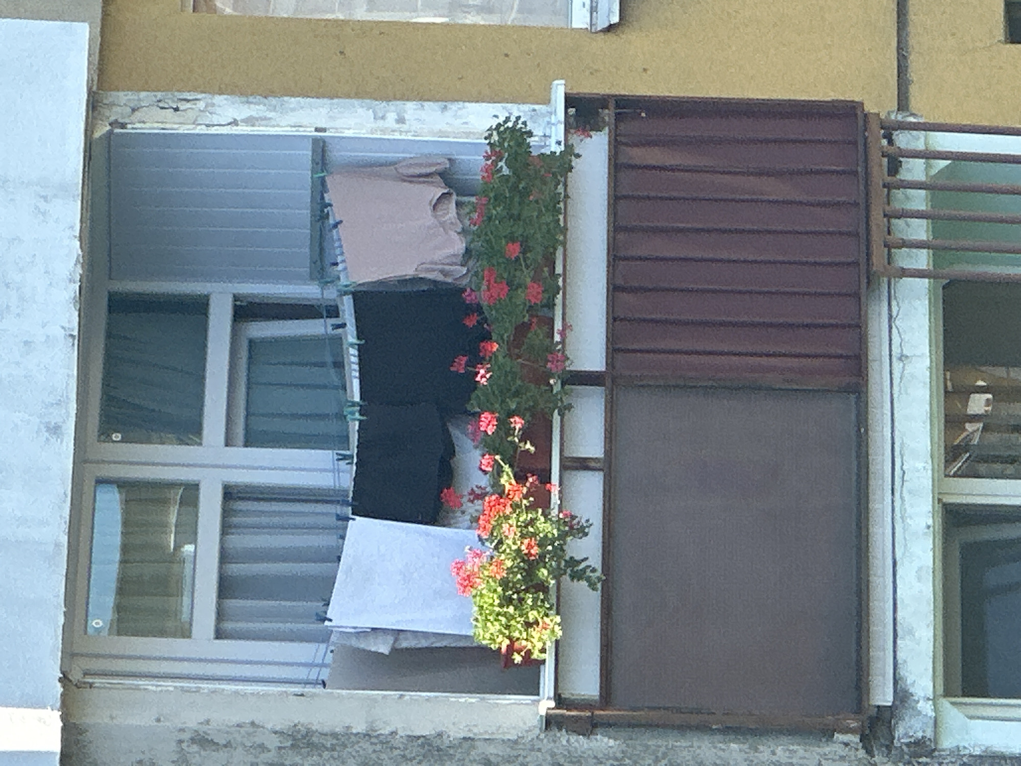 This image depicts a balcony on a multi-story building in Uzhhorod, Ukraine. The balcony has a window with a light blue-tinted glass and vertical blinds. Laundry is hanging on a clothesline across the window, including a white sheet, dark fabric, and a light pink garment. Flower pots with red flowers and green foliage are arranged on the windowsill. Below the window, the balcony railing consists of brown metal panels and vertical bars. The building exterior shows signs of weathering, with peeling paint and concrete. In the background, another building with a yellow facade is visible.