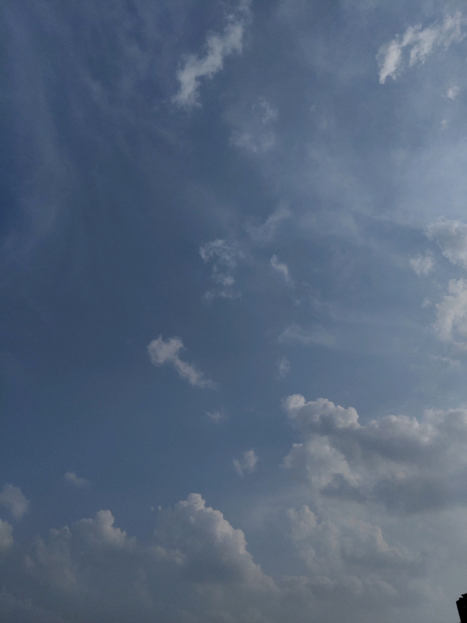 A photograph captures a view of a cloudy sky over Fajar Baru, Indonesia. The sky is a mix of light and dark blues, with various cloud formations scattered across it. Some clouds are wispy and thin, appearing like streaks across the sky, while others are thicker and more defined, resembling cotton balls. In the lower right corner of the image, a dark, silhouetted object, likely a building, is partially visible against the sky.