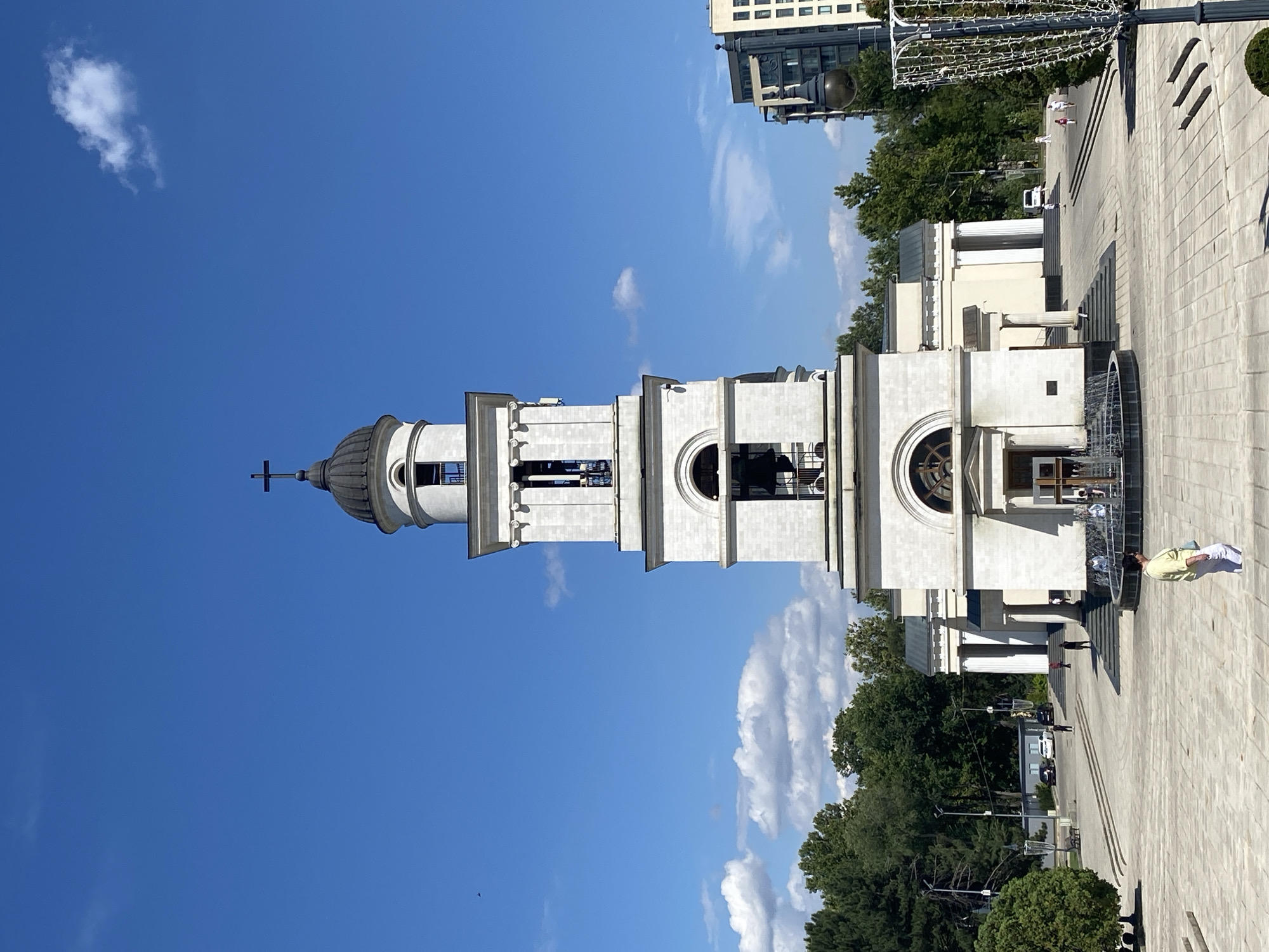 The image depicts a wide, sunlit plaza in front of the Cathedral of Christ's Nativity in Chișinău, Moldova. The prominent feature is the cathedral's bell tower, a white, multi-tiered structure topped with a dark dome and a cross. The tower has arched openings revealing bells within and is flanked by the main body of the church, characterized by columns and a pedimented entrance.
The plaza itself is paved with light-colored stone tiles and features a circular fountain in the foreground. Several individuals are visible: a woman in a yellow top and white pants walks away from the viewer in the foreground. Other people are scattered around the fountain area, near the church entrance, and in the middle distance, some walking and others appearing to gather.
Behind the cathedral, a line of lush green trees forms a backdrop. To the right of the scene, modern apartment buildings are visible, contrasting with the historical architecture of the cathedral. The sky is a clear, bright blue with scattered white cumulus clouds. The lighting suggests it is midday. The overall atmosphere is peaceful and public, capturing a moment of everyday life in a significant city square.