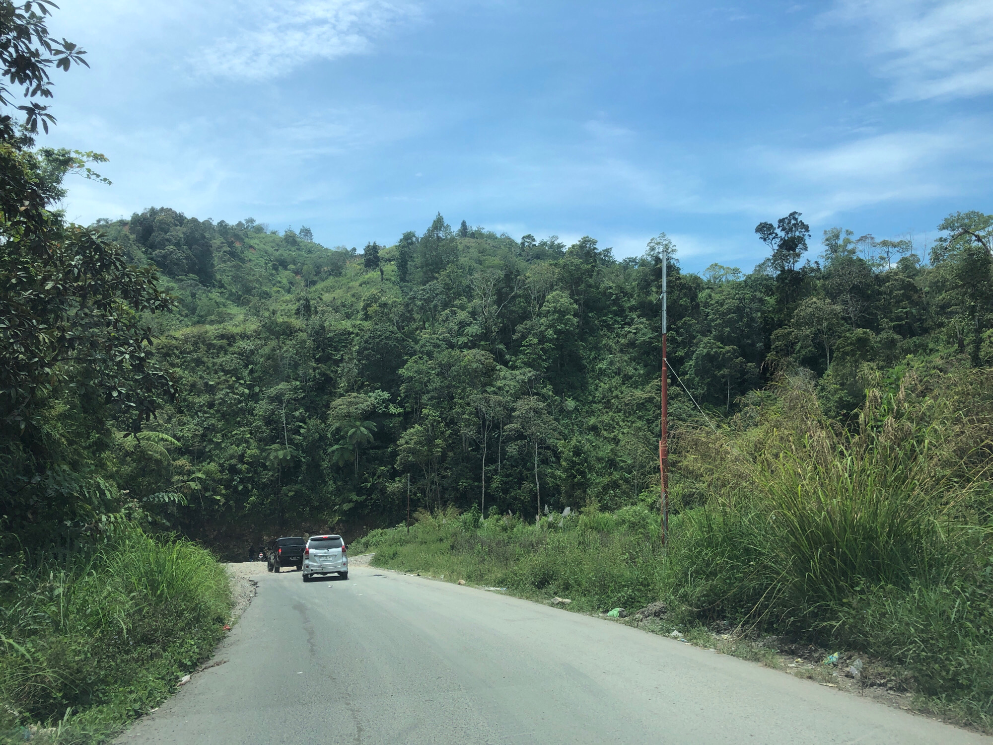 The image depicts a two-lane asphalt road winding through a lush, green, mountainous landscape. The road is bordered by dense vegetation, including tall grasses and trees that climb the hillsides. The sky is partly cloudy with a bright blue hue. Two vehicles are visible on the road; a silver SUV is in the foreground, and a black vehicle is further ahead. The scene appears to be a rural or natural setting, possibly a scenic route. The location is identified as Bintangpermata, Indonesia.