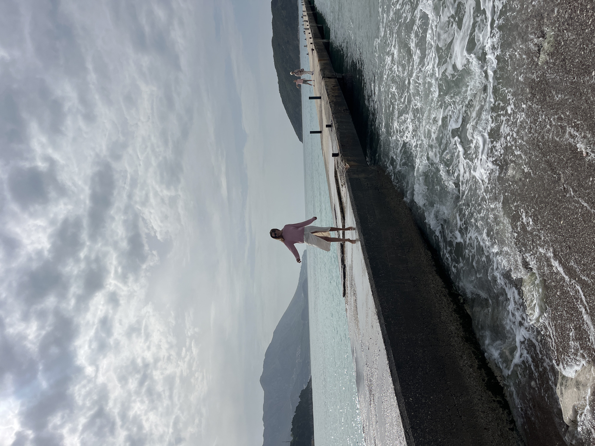 A person in a pink shirt and white skirt stands with outstretched arms on a concrete pier next to choppy water. Another figure is visible further down the pier. A mountain and cloudy sky are in the background. The location is likely Budva, Montenegro.