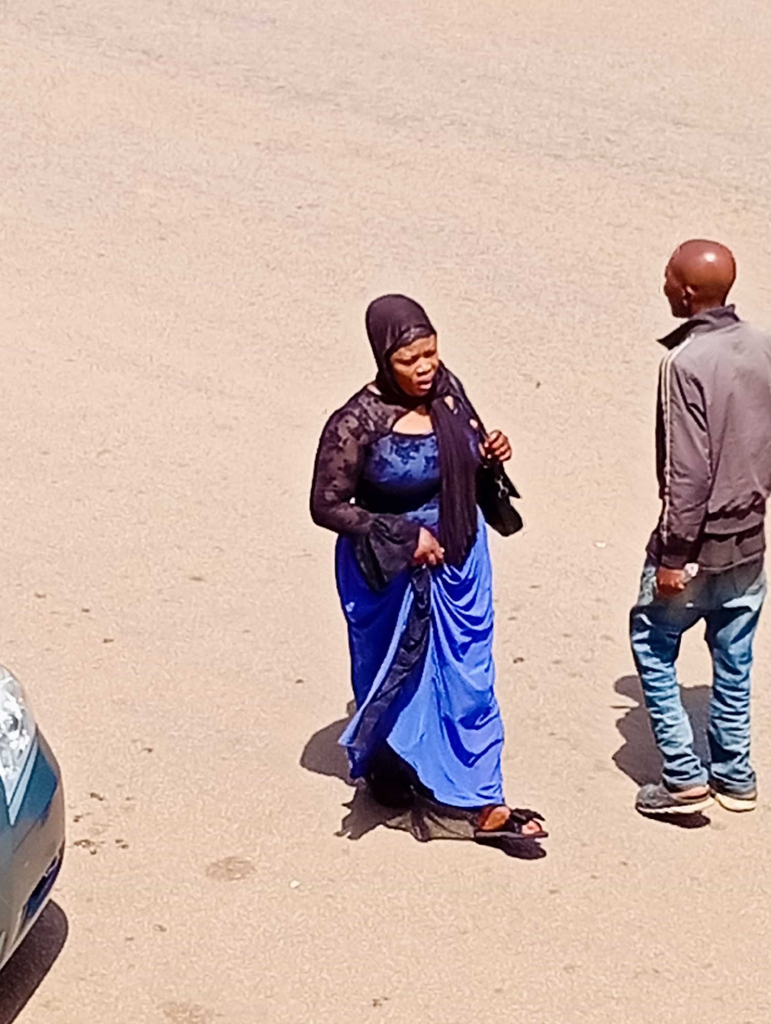 In Bukuru, Nigeria, two individuals are standing on a paved surface. On the left, a person wearing a black hijab and a blue dress with black lace detailing is looking towards the right. They are holding a dark-colored bag in their right hand. On the right, a person with a shaved head, wearing a grey jacket and blue jeans, is facing away from the camera and towards the other individual. The ground is a light brown, sandy texture, with a car partially visible on the far left of the frame. Shadows are cast on the ground, indicating a sunny day.