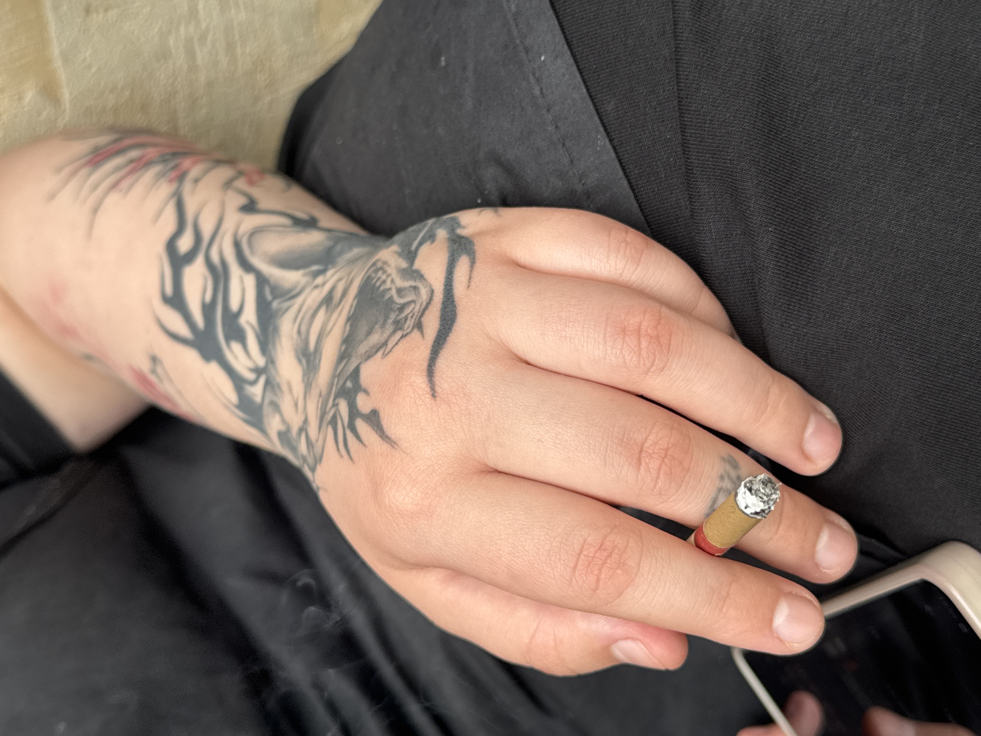 The media file is a close-up shot of a person's hand and arm, with a cigarette held between the fingers. The person has an intricate tattoo covering their arm, featuring what appears to be a stylized tiger or dragon. The cigarette is lit, with ash at the tip, and a wisp of smoke is visible, suggesting it is being smoked. The person is wearing a black garment, possibly a t-shirt or jacket. A portion of a light-colored phone case is visible in the lower right corner of the frame. The lighting suggests it is daytime, and the background is blurred, focusing attention on the hand and arm. The setting appears to be indoors or in a shaded area. No text is visible in the image. The overall impression is candid and focuses on the detail of the tattoo and the act of smoking.