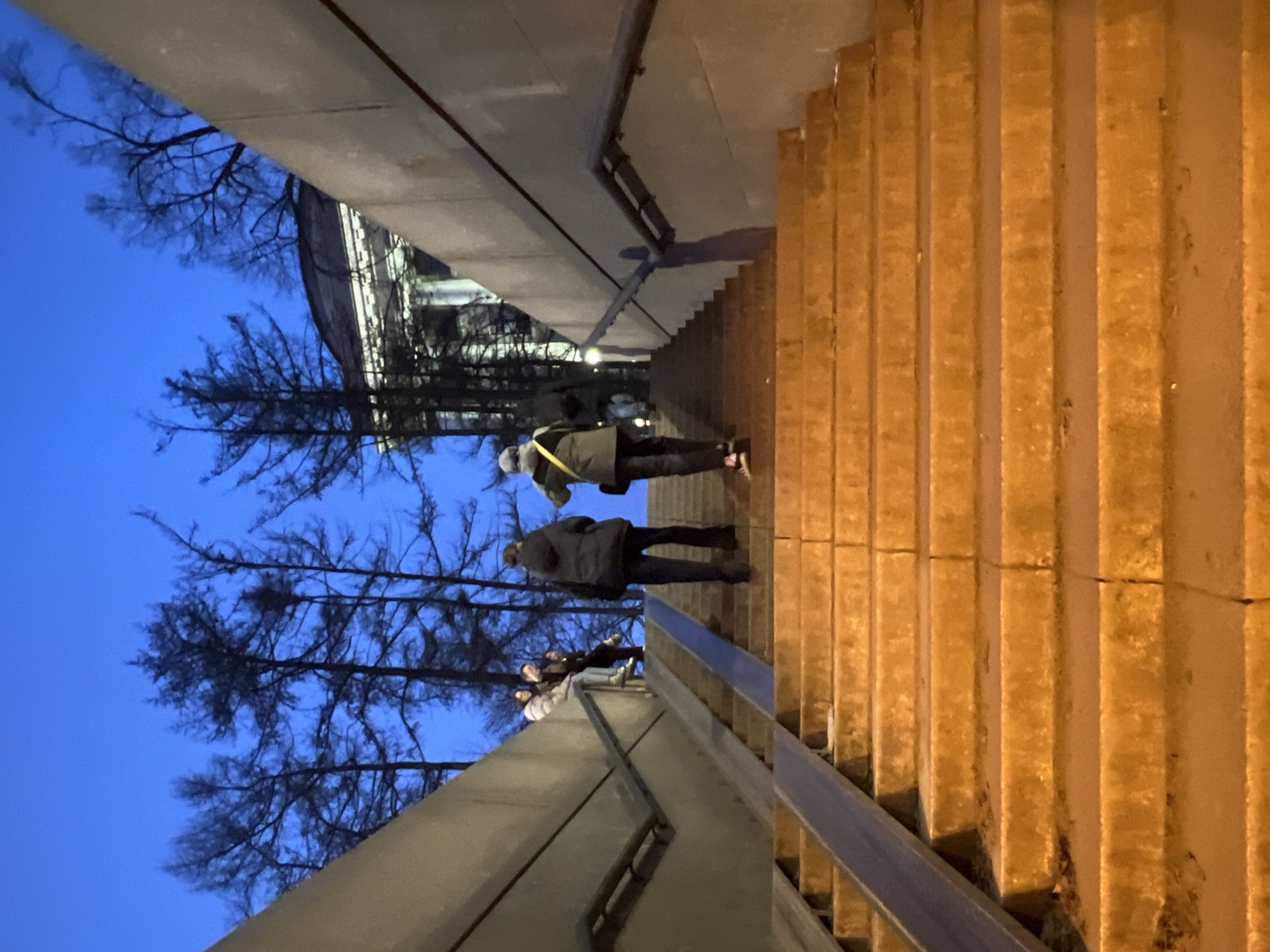 The image depicts an urban pedestrian stairwell in Saint Petersburg, Russia, at dusk or early evening, likely an exit from an underpass or metro station leading towards the Alexander Garden area, with a visible portion of the classical Admiralty Building in the background. The concrete staircase, appearing worn, ascends between two high, unadorned concrete walls, illuminated by a warm, orange artificial light. Dark metal handrails are affixed to both walls. A dark-colored metal ramp for wheeled transport runs parallel to the stairs on the left. In the immediate foreground, two individuals are actively ascending the stairs, viewed from behind: one wears a dark puffer jacket and dark trousers, while the other wears an olive-green jacket with a yellow strap visible across the back, dark trousers, and a light-colored beanie. Higher on the left side of the staircase, a group of three individuals is seated on a concrete ledge or step. Additional indistinct figures are visible further up the stairs on the right. Above the stairwell structure, the silhouettes of several leafless trees are starkly contrasted against a deep blue twilight sky. A white, classical building with visible columns and a domed or rounded roof structure is partially discernible through the trees on the upper right.