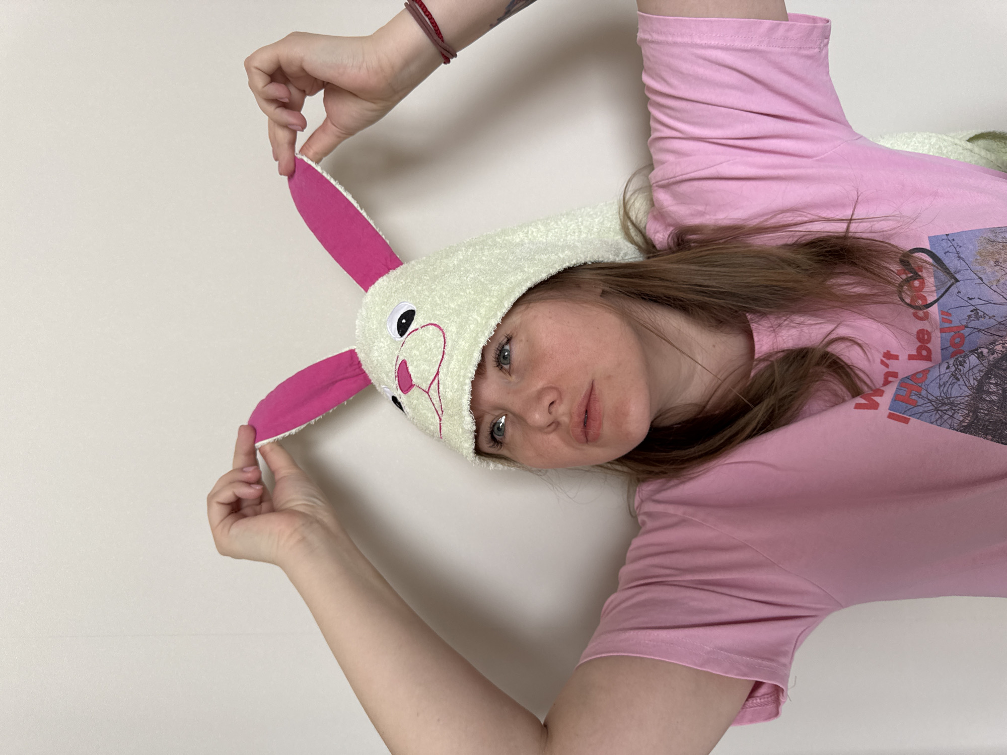 A Caucasian female, estimated to be in her twenties or thirties, is depicted indoors against a plain, light beige wall in Gornyi (Dvurechenskoe S/P), Russia. She is wearing a light pink, short-sleeved t-shirt with a partially visible graphic print on the chest, and a light green, fuzzy rabbit-themed hat featuring pink inner ears, black eyes, and a pink nose/mouth embroidery. Her arms are raised, with both hands grasping the tips of the rabbit ears on her hat. Her head is tilted slightly upwards and to her right, with her blue eyes looking in the same direction. Her mouth is slightly open. Several thin bracelets, including one red and one brownish-red, are visible on her left wrist, and a dark tattoo is partially visible on her left forearm.