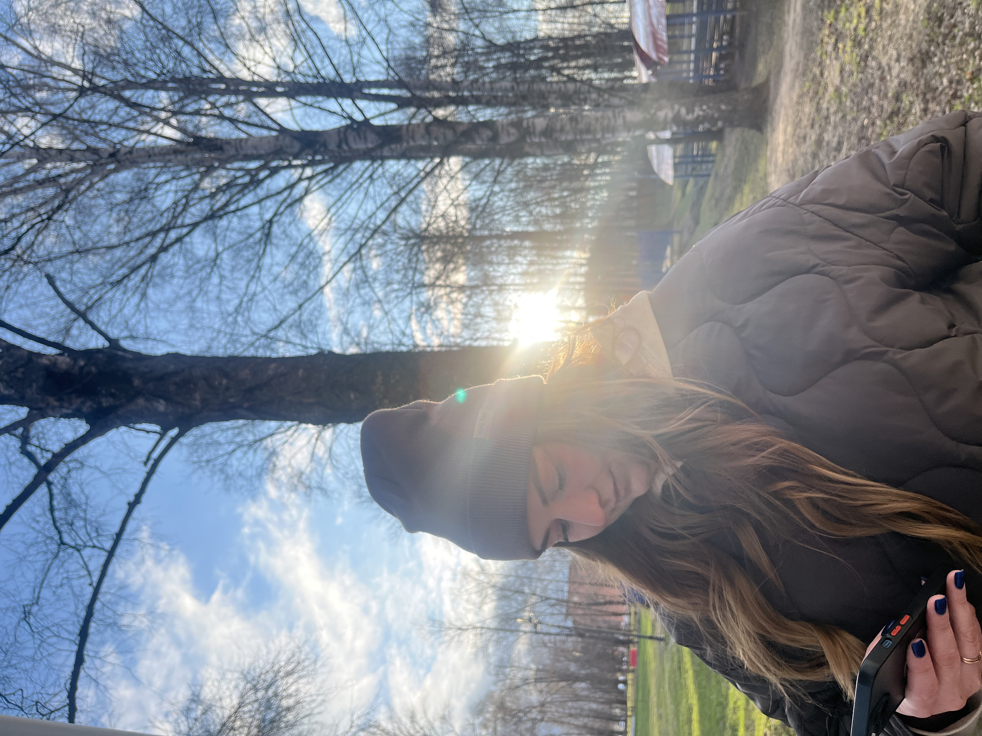 A woman in a brown puffer jacket and a gray beanie is looking at a black smartphone in her right hand. The background includes bare trees against a blue sky with white clouds, sunlight, and green grass. The location is Klin, Russia.