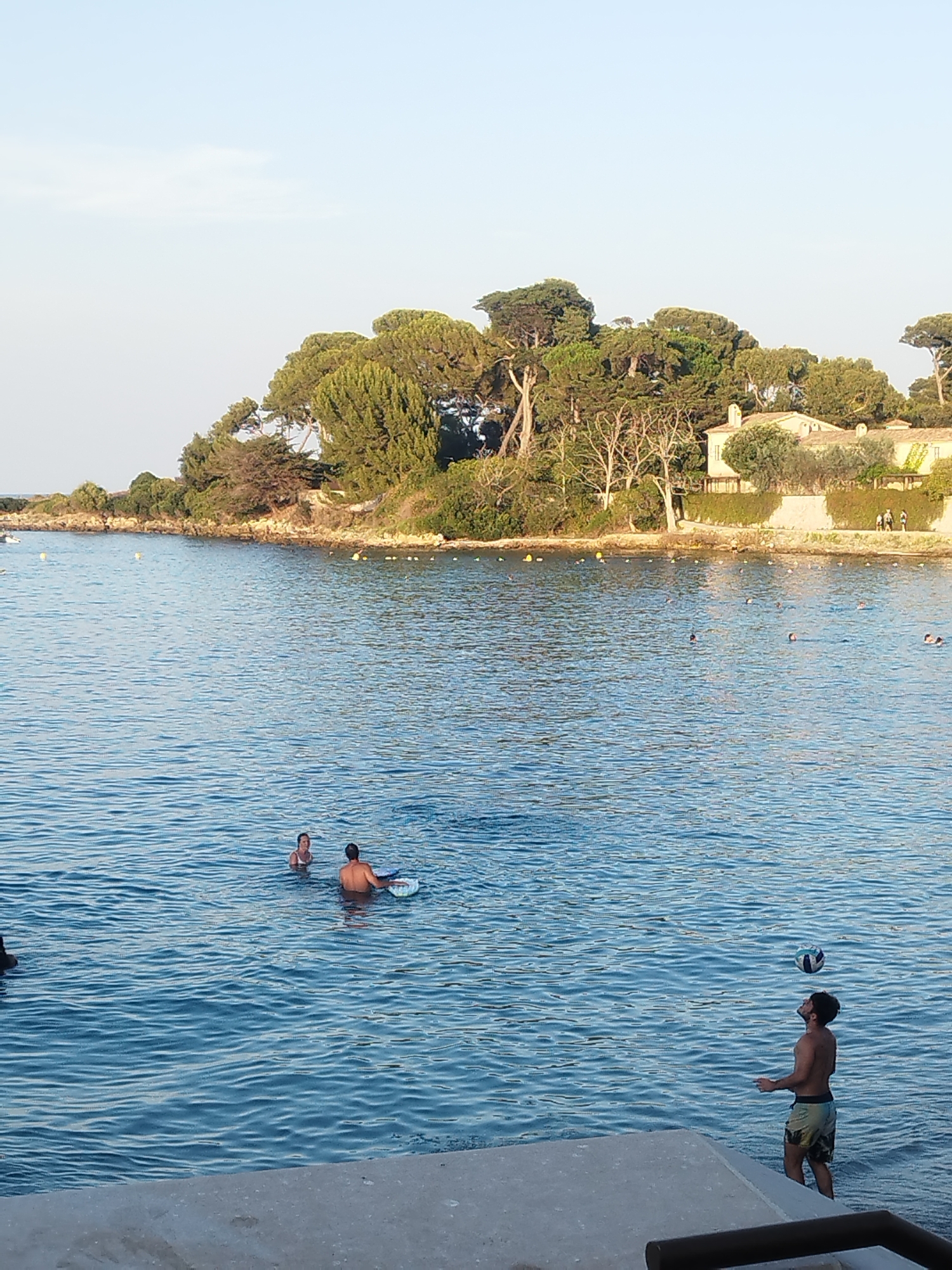 The image depicts a waterfront scene in Antibes, France, likely during a sunny day, based on the clear sky and water. In the foreground, a person stands partially in the water near a concrete edge, looking up at a volleyball they are about to serve or have just served. Further out in the water, two people are visible, one on a float and another standing beside them. The background features a tree-lined shore with lush green foliage and a building with a pale yellow facade. The sea is calm with gentle ripples.