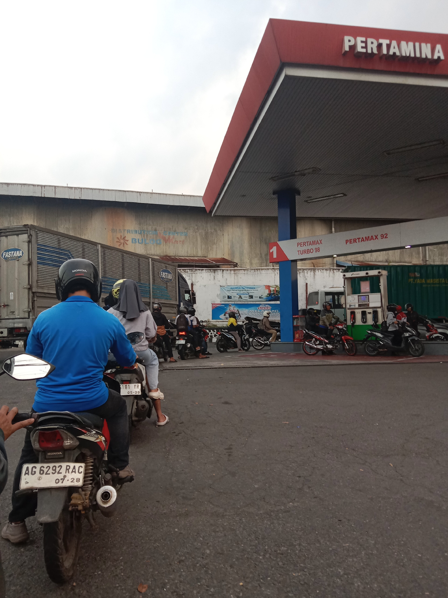 A queue of motorcyclists waits at a Pertamina gas station in Semarang, Indonesia. The foreground features a motorcycle with a rider wearing a blue shirt and black helmet, and a passenger in a grey hijab. Behind them, other motorcyclists are lined up. To the right, a gas pump island is visible with several vehicles. In the background, a large truck with "FASTANA LOGISTICS" and "PSP" branding is parked. The Pertamina signage is prominently displayed on the canopy of the gas station. The scene is set against a cloudy sky and appears to be a typical day at a busy urban fuel station.