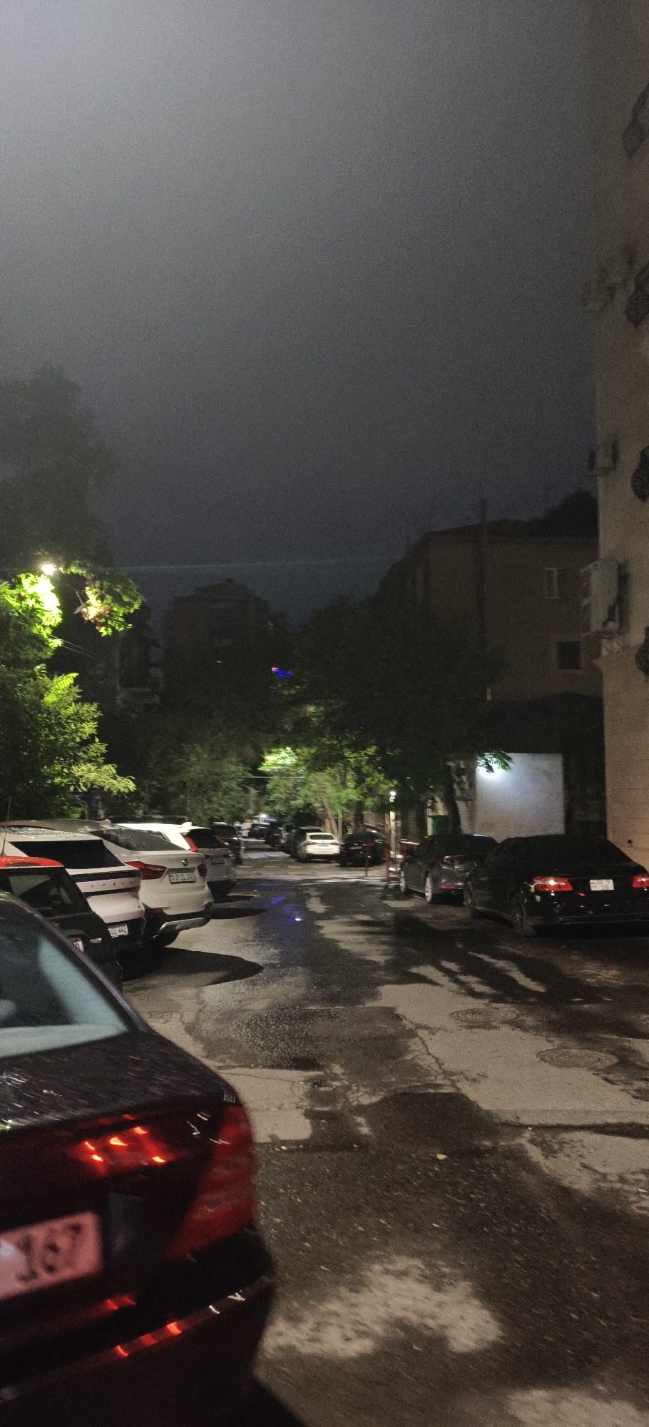 A nighttime scene depicts a street lined with parked cars in an urban environment. The road surface appears wet, suggesting recent rain or mist. In the foreground, the rear of a dark-colored sedan is visible, with its taillights illuminated. To the left, several other vehicles are parked, including a white SUV. Trees with green foliage are present along the street, some illuminated by overhead lights. In the background, buildings with visible windows can be seen, and the sky is dark and obscured by what appears to be fog or low cloud cover. The overall atmosphere is dim and somewhat hazy.