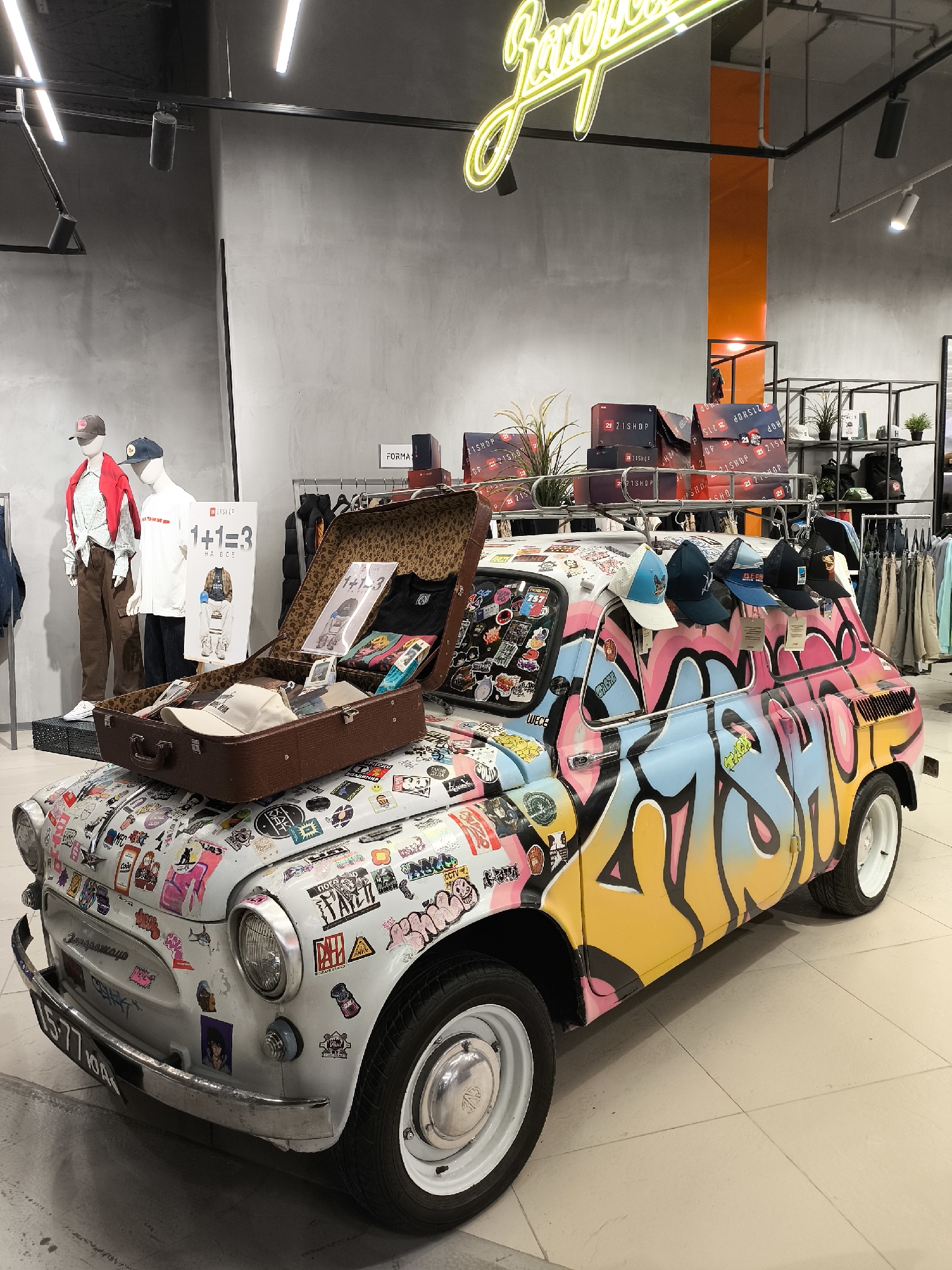 This indoor scene features a sticker-covered and graffiti-painted car as a display inside a store. The car is a vintage model, with a suitcase opened on its hood revealing clothing items and signage. A collection of hats hangs from the car's roof. The car's license plate reads "A577 YOA". In the background, two mannequins are dressed in casual clothing. Clothing racks and stacked boxes bearing the "21SHOP" logo are visible, along with a neon sign. The store's walls are a neutral gray, and lighting is provided by overhead fixtures.