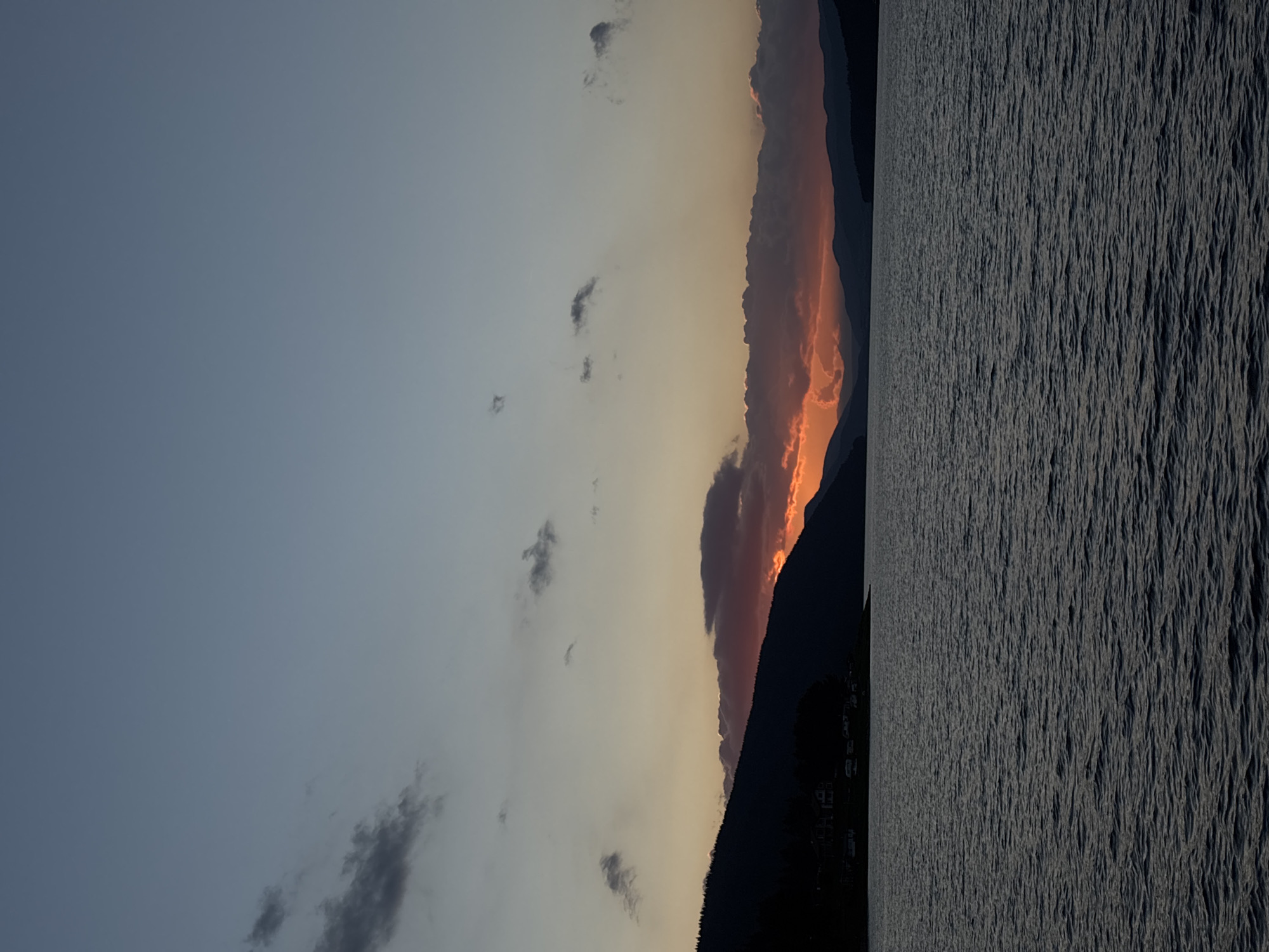 The image depicts a body of water, likely a lake or ocean, at dusk. The water's surface is choppy, reflecting the dim light of the sky. Silhouetted against the horizon are mountains and a forested shoreline. The sky is a gradient of colors, transitioning from a pale blue at the top to shades of orange and pink near the horizon, indicating the setting sun. Scattered clouds are visible in the sky, some dark and others illuminated by the last rays of sunlight. The overall atmosphere is serene and tranquil.